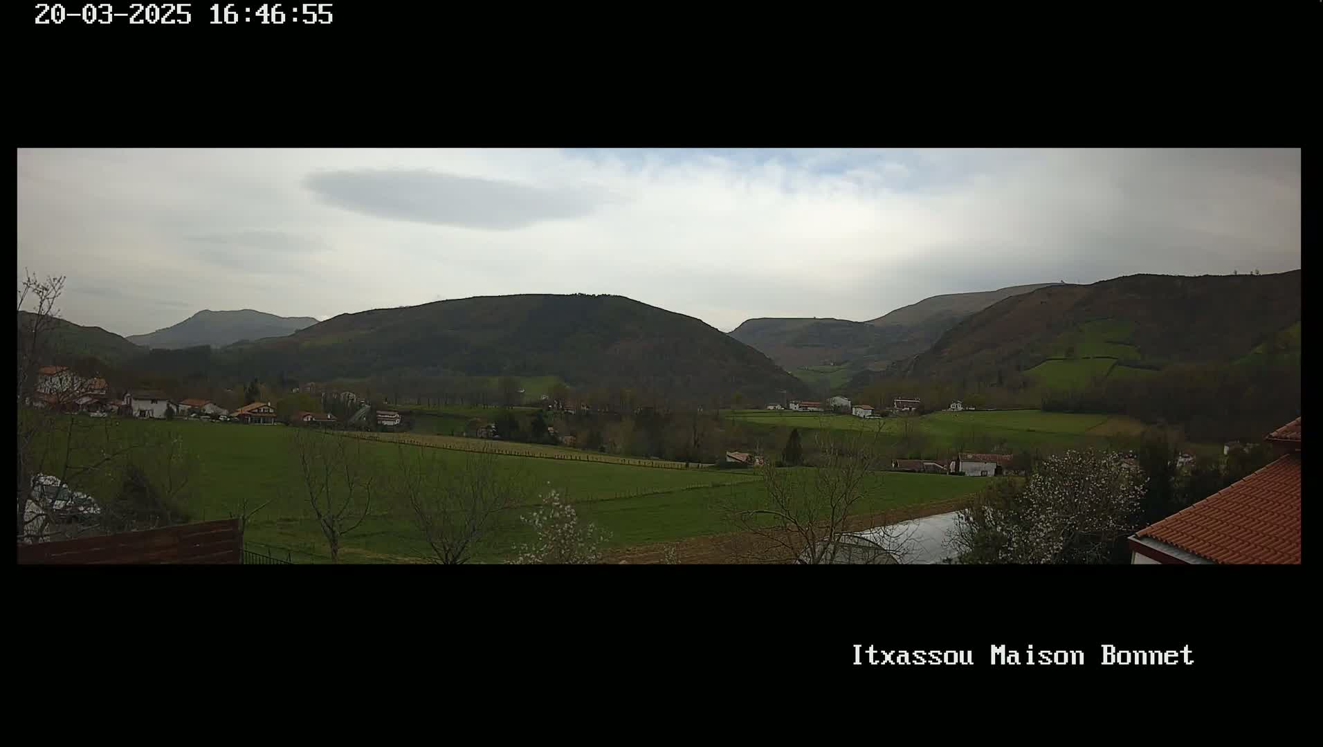 A panoramic view of a rural landscape under an overcast sky shows rolling green hills, farmlands, and a few scattered houses.