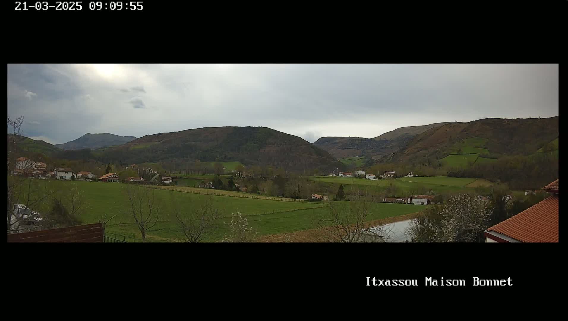 A panoramic view of a rural valley with overcast skies shows green fields, scattered houses, and rolling hills.