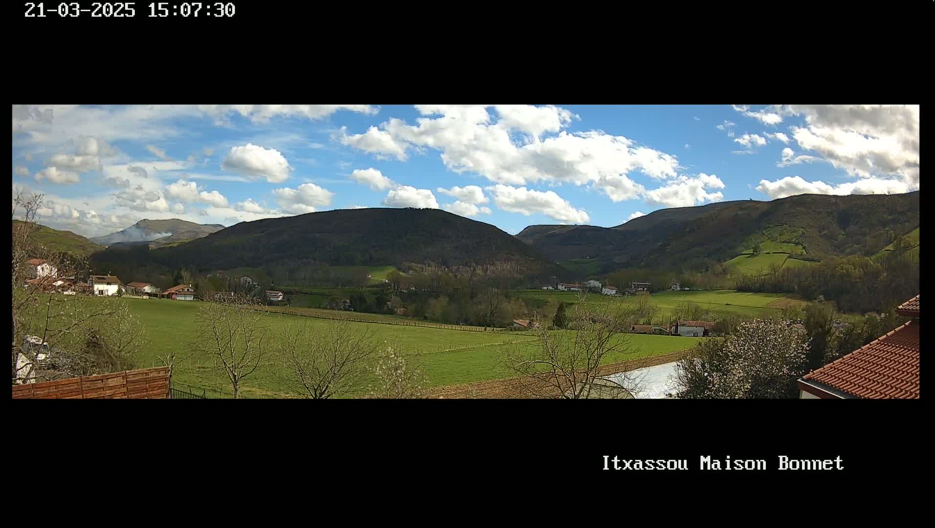 A partly cloudy day reveals a verdant valley nestled between rolling hills, dotted with small buildings and farmlands.