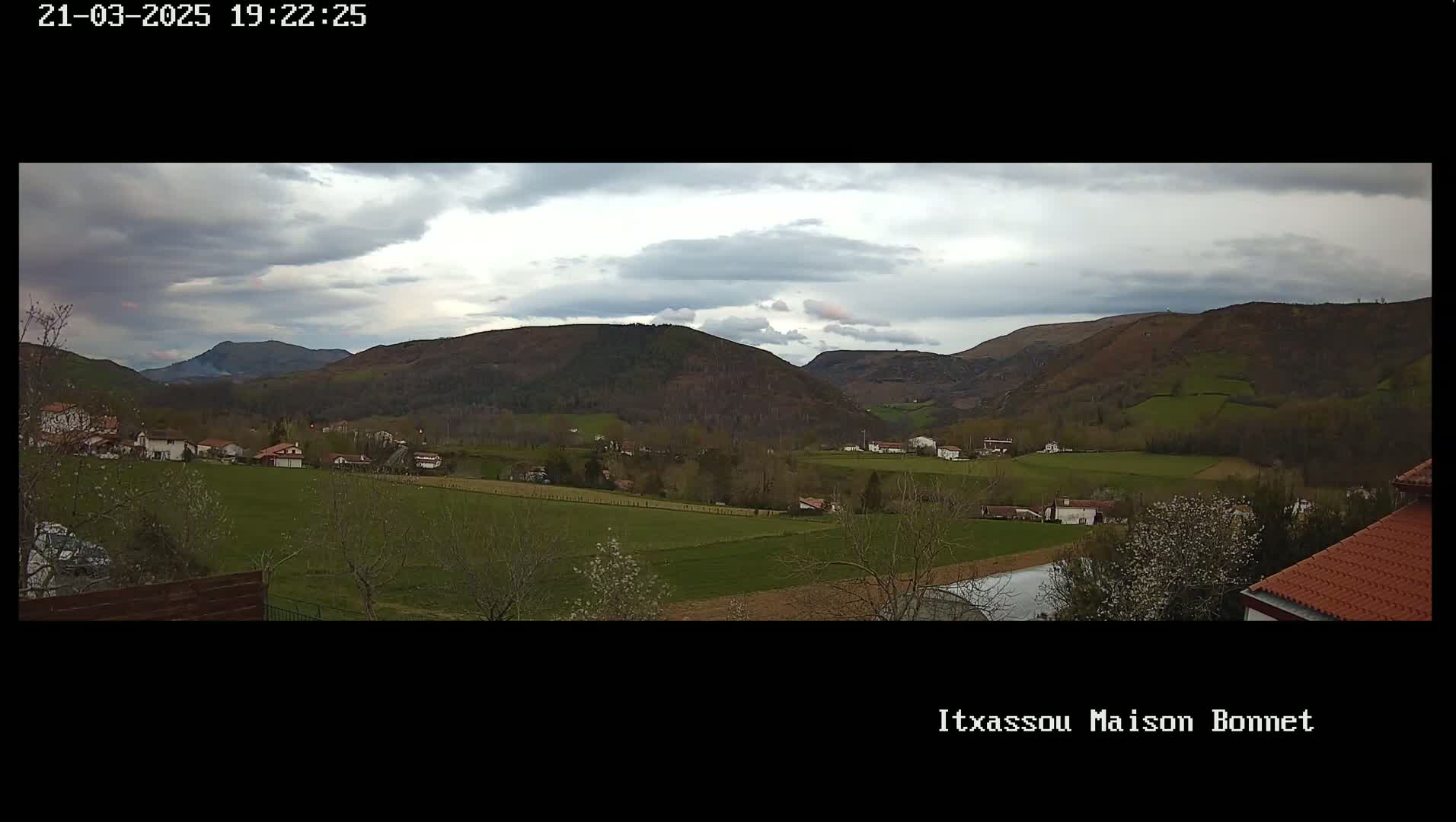 A panoramic view of a rural valley under a cloudy sky shows a scattering of houses and fields nestled among rolling green hills.