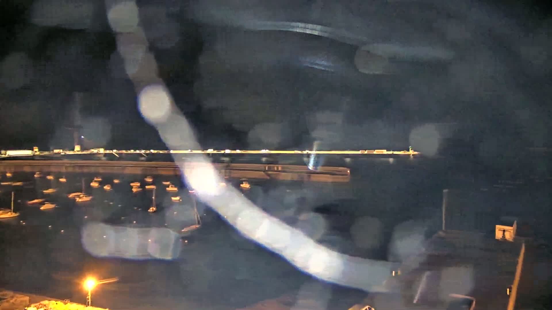A nighttime view of a harbor filled with boats, seen through rain-covered glass, with a distant shoreline illuminated.