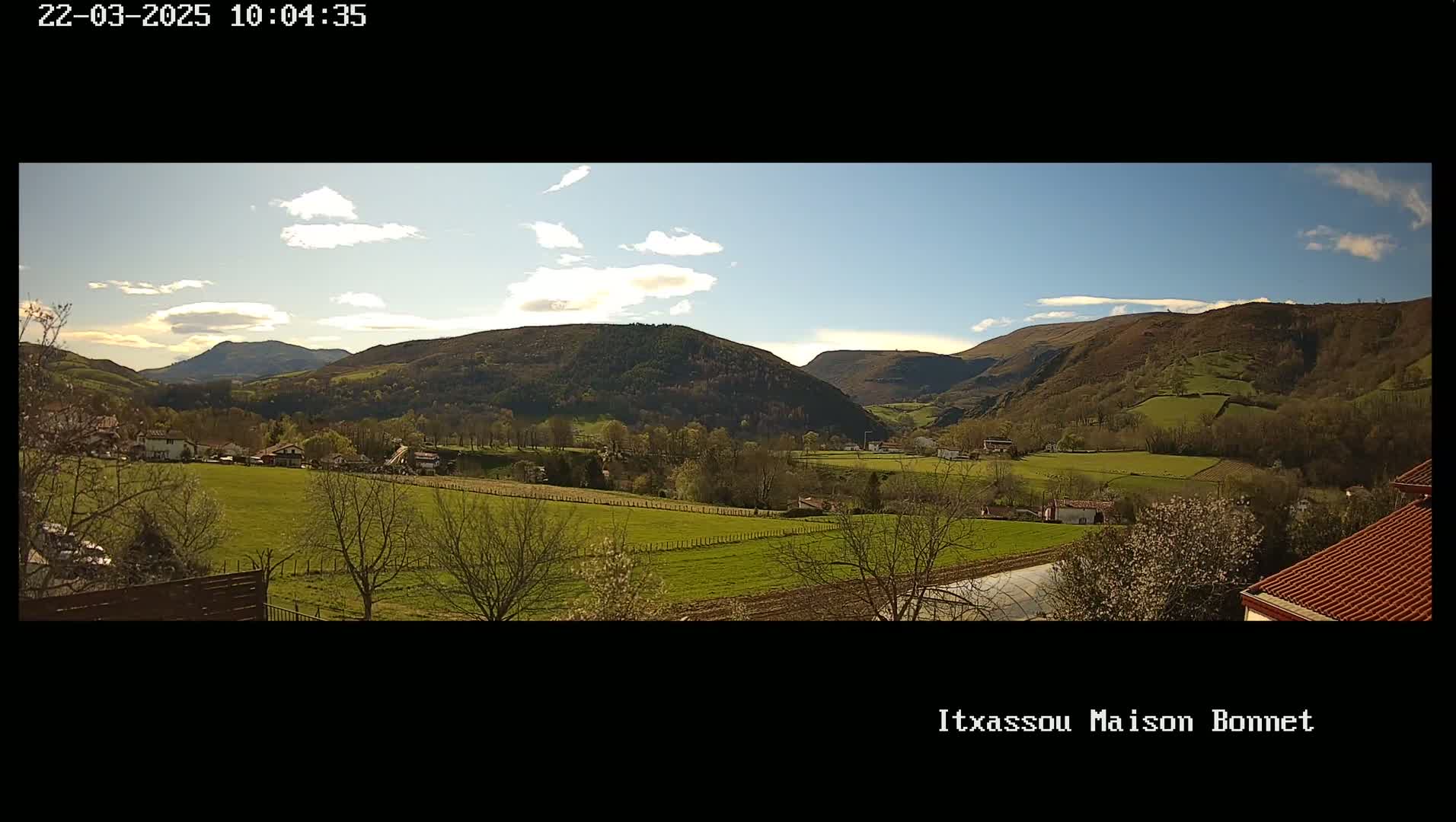 A sunny day reveals a valley of green fields and trees, nestled between rolling hills under a partly cloudy sky.