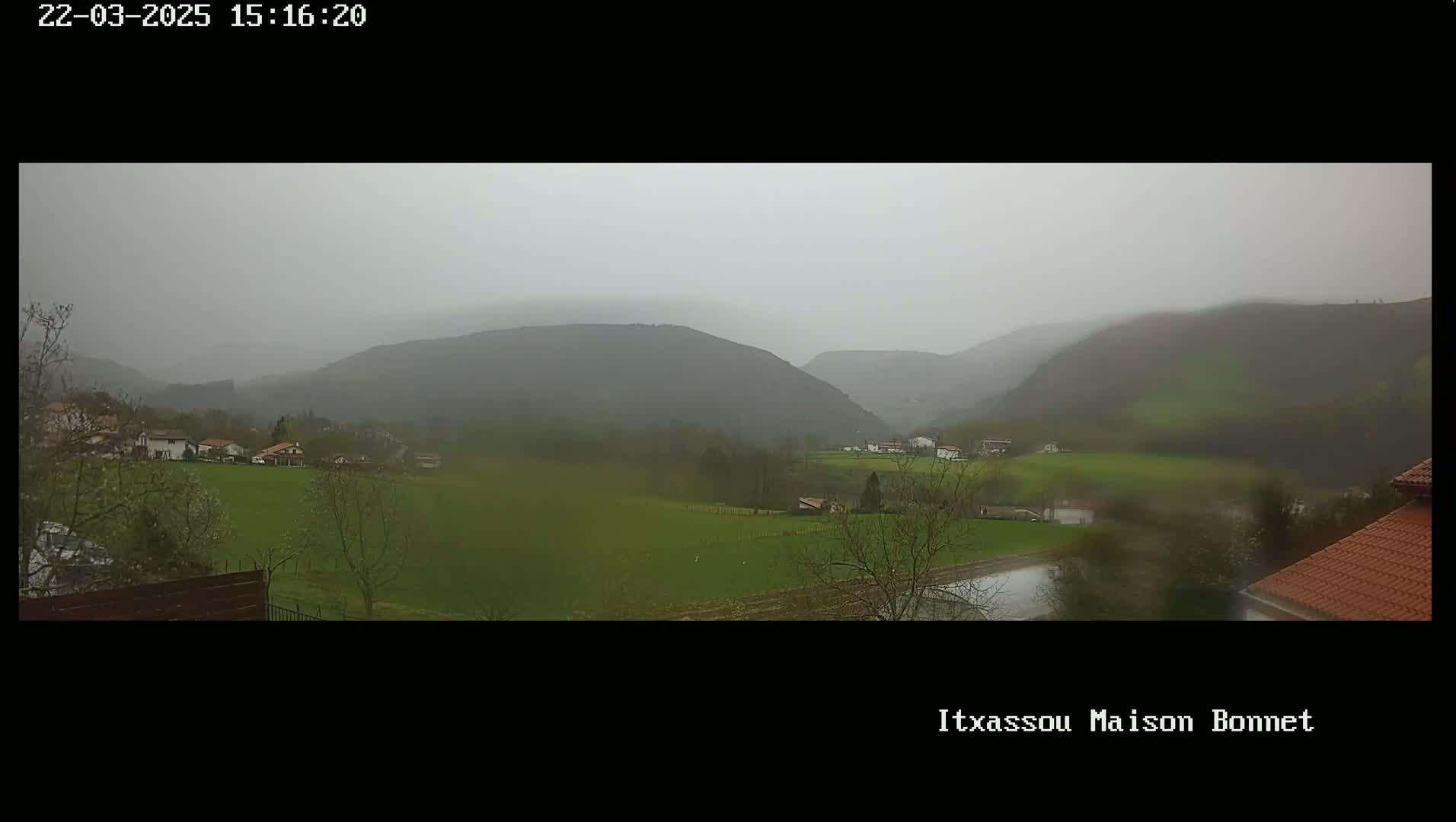 A view of a valley with rolling green hills and a few houses under an overcast, rainy sky.