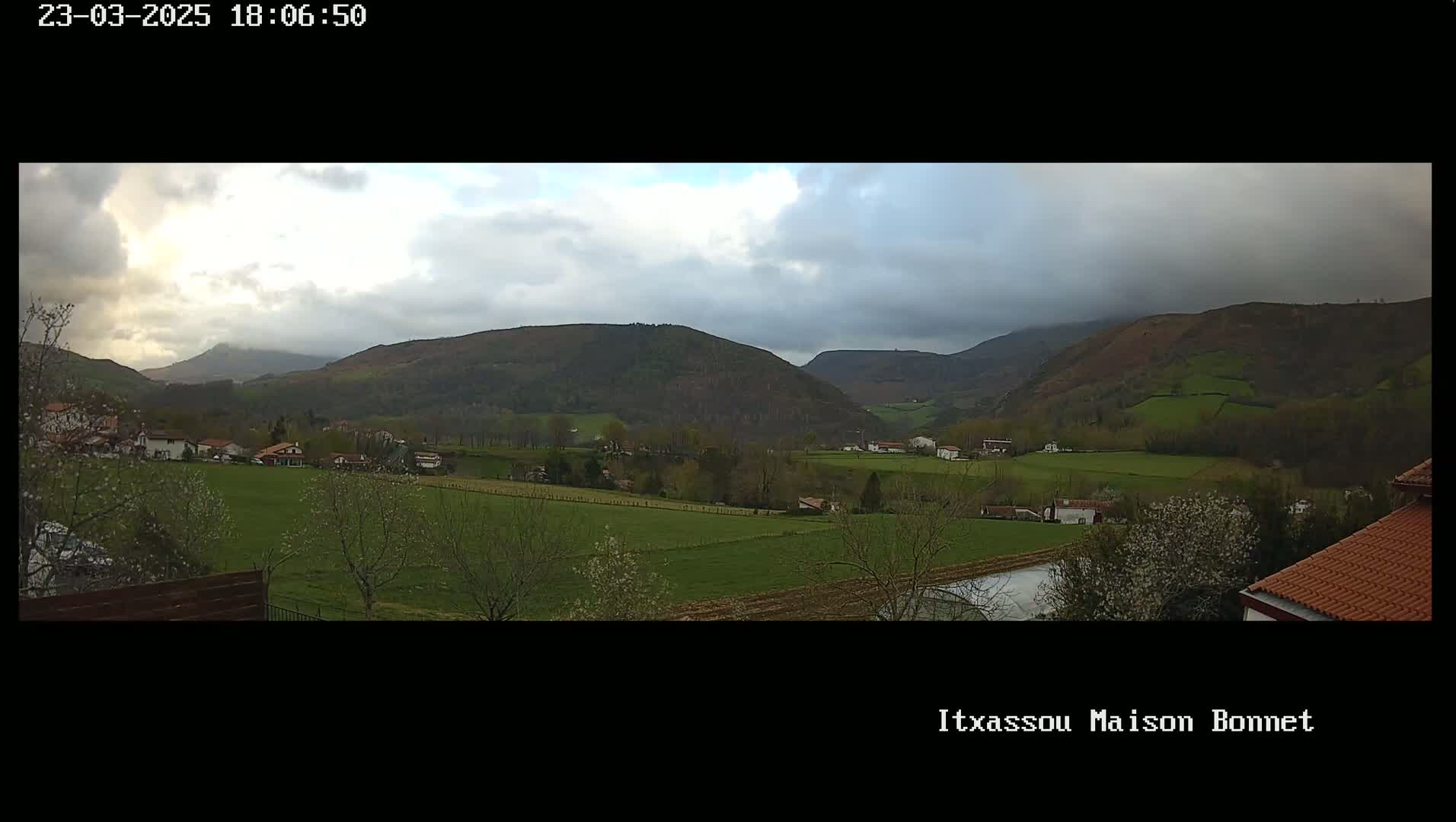 A mostly cloudy day reveals a sprawling valley dotted with houses and farmland, nestled between gently rolling green hills.