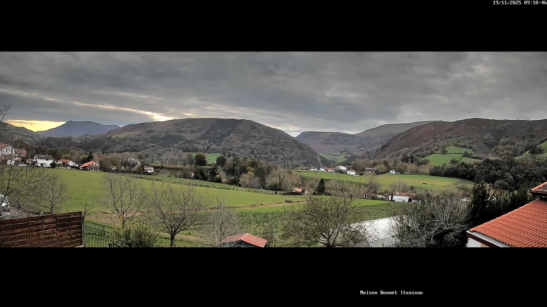 A panoramic view captures a verdant valley dotted with scattered homes and a mix of bare and forested trees, nestled among rolling hills under a heavily overcast sky with lighter clouds breaking through in the distance.