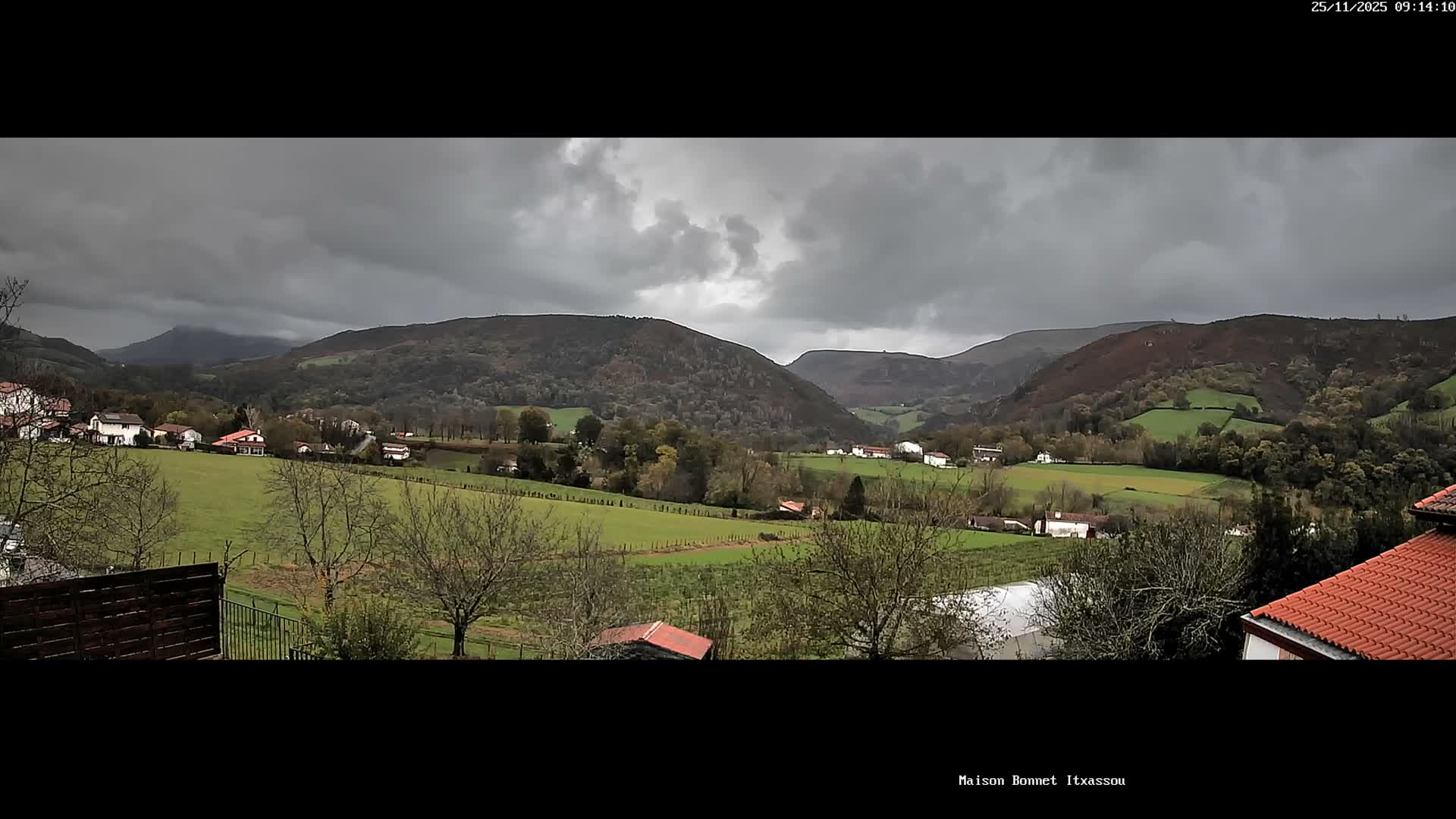 A wide panoramic view captures a rural valley dotted with houses and green fields surrounded by tree-covered hills under a heavily overcast and gloomy sky.
