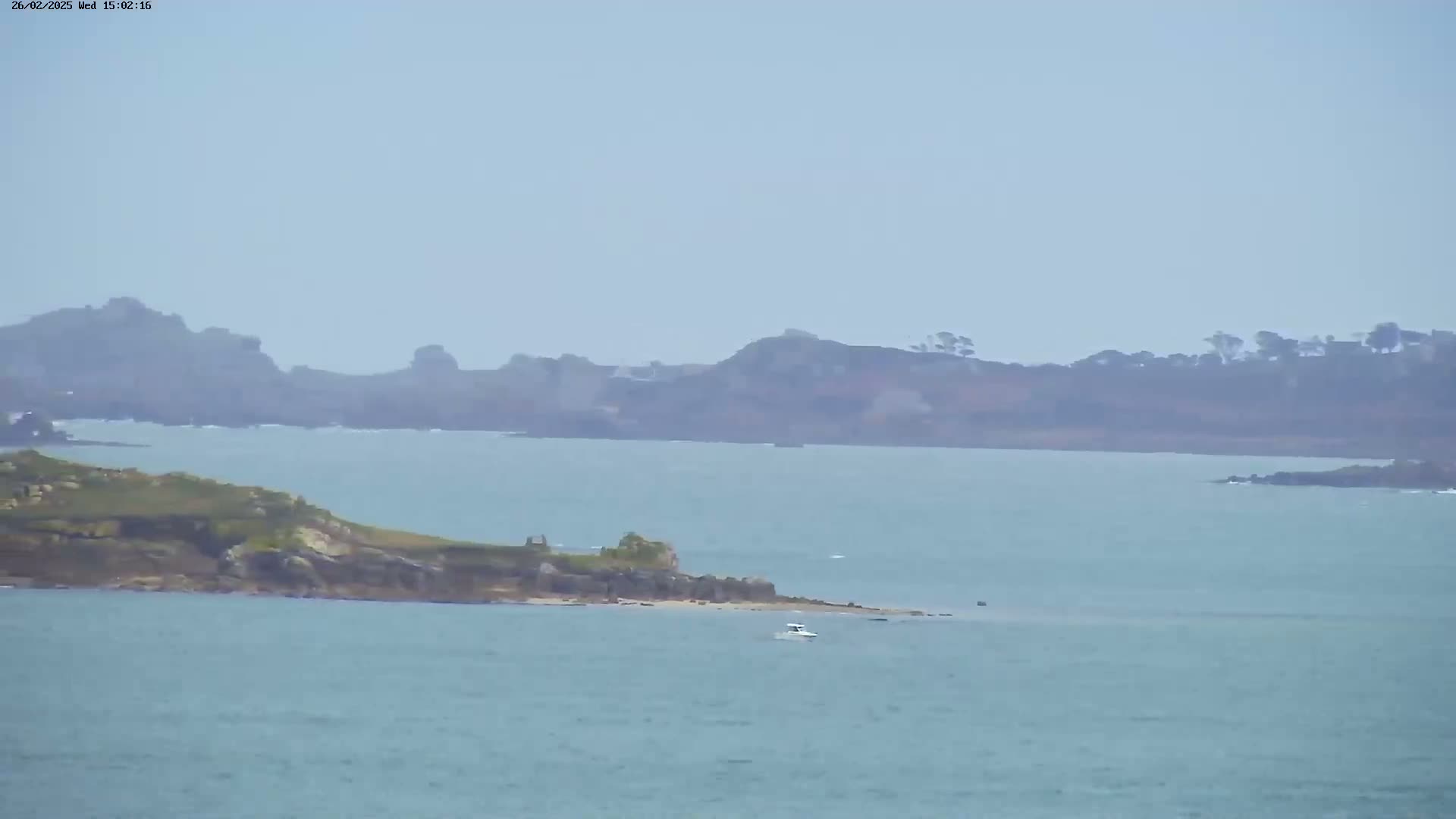 A hazy view shows a small boat on calm water near a small, rocky island, with larger landmasses visible in the distance.