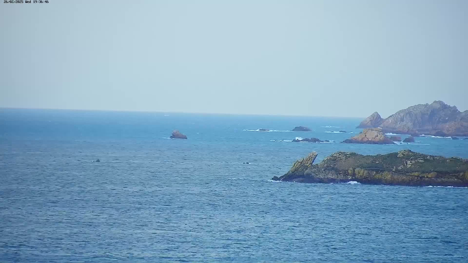 A rocky outcrop and several other small islands are visible in a calm, blue ocean under a hazy sky.