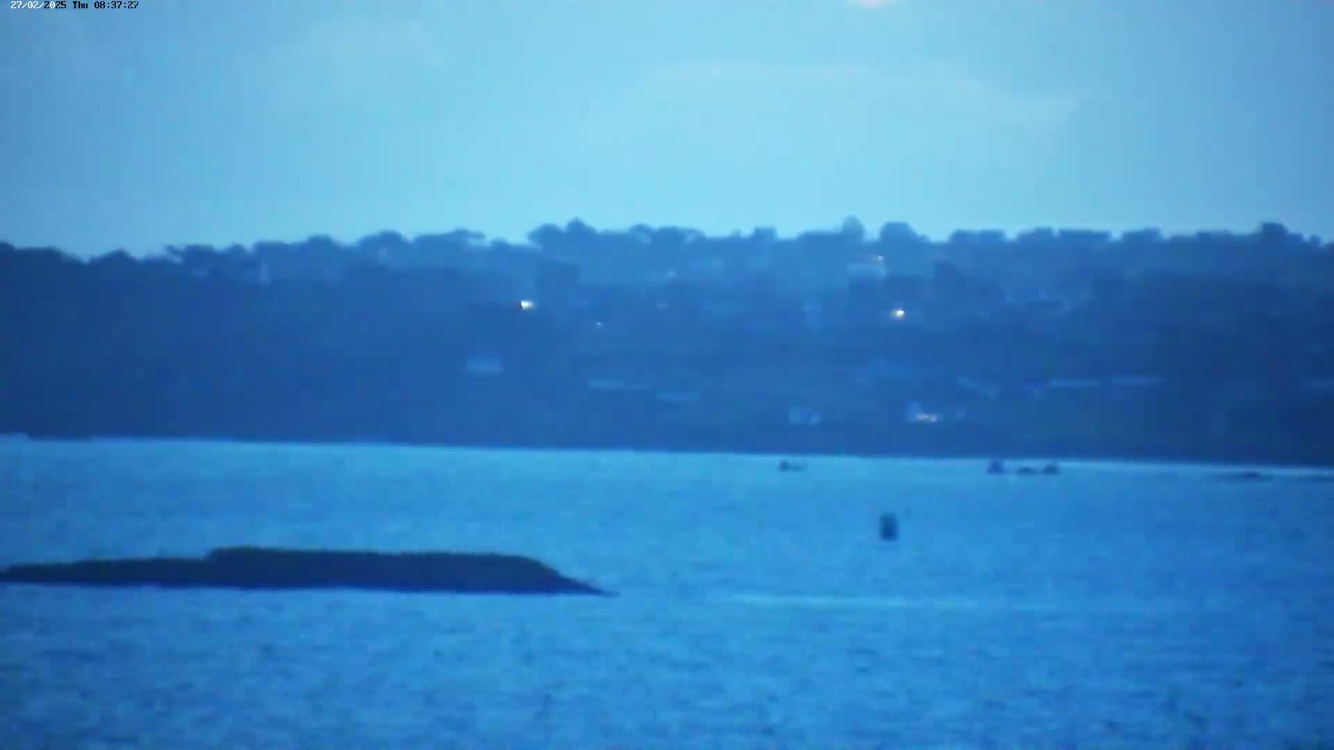 A hazy, calm body of water shows a distant shoreline with a town visible, and a small, dark rock formation closer to the viewer in the foreground.