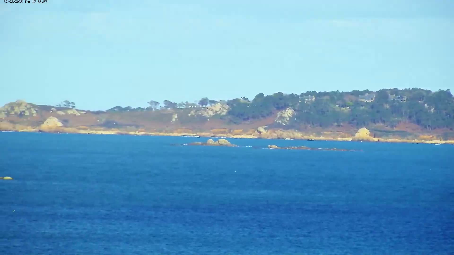 A calm, blue ocean stretches to a distant shoreline of rocky coast and dark green trees under a clear, light blue sky.