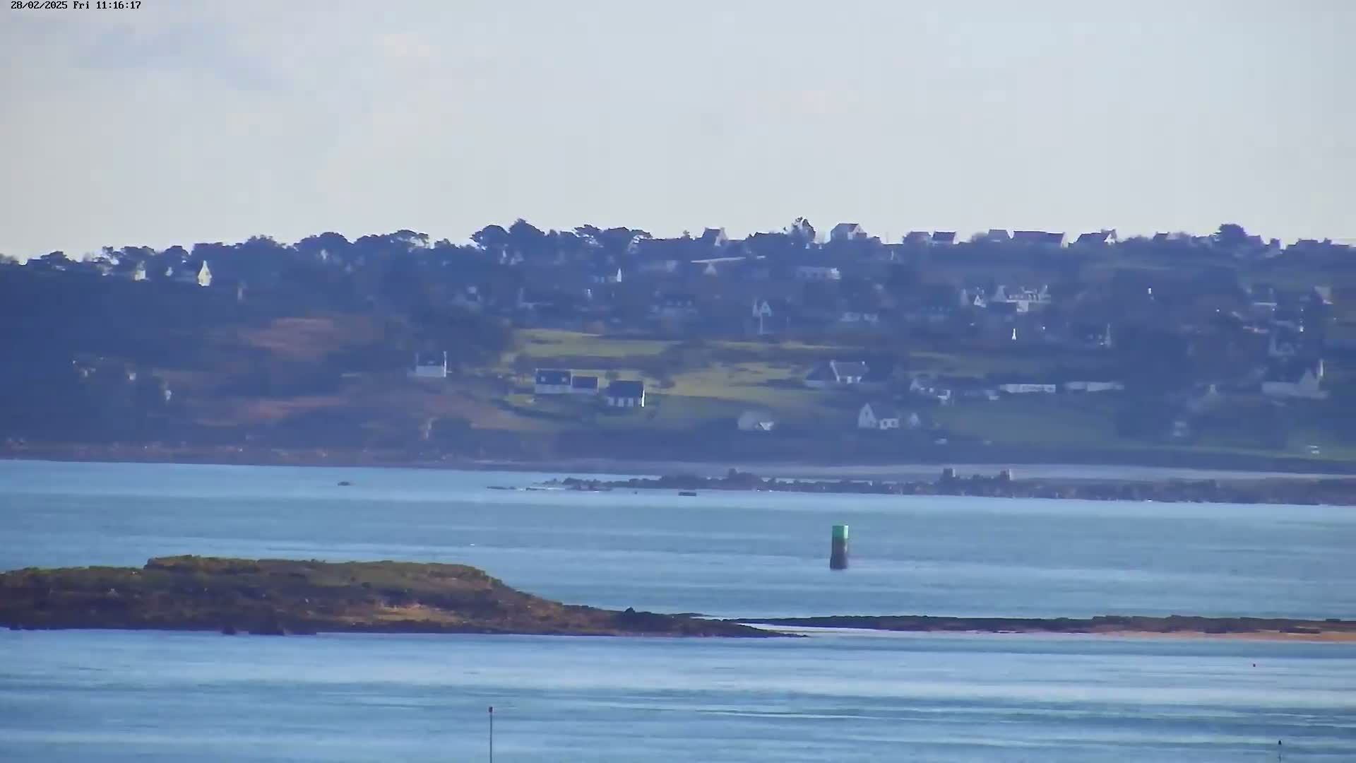 A coastal scene on a hazy day shows a small island in the foreground, a calm body of water, and a distant village nestled on a hillside.