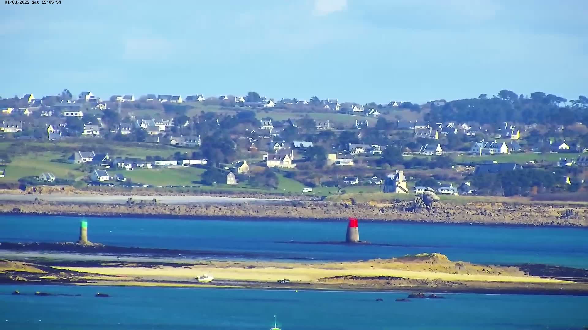 A coastal town is visible in the distance across a body of blue water, under a clear sky; two small, colored markers are situated in the water in the foreground.