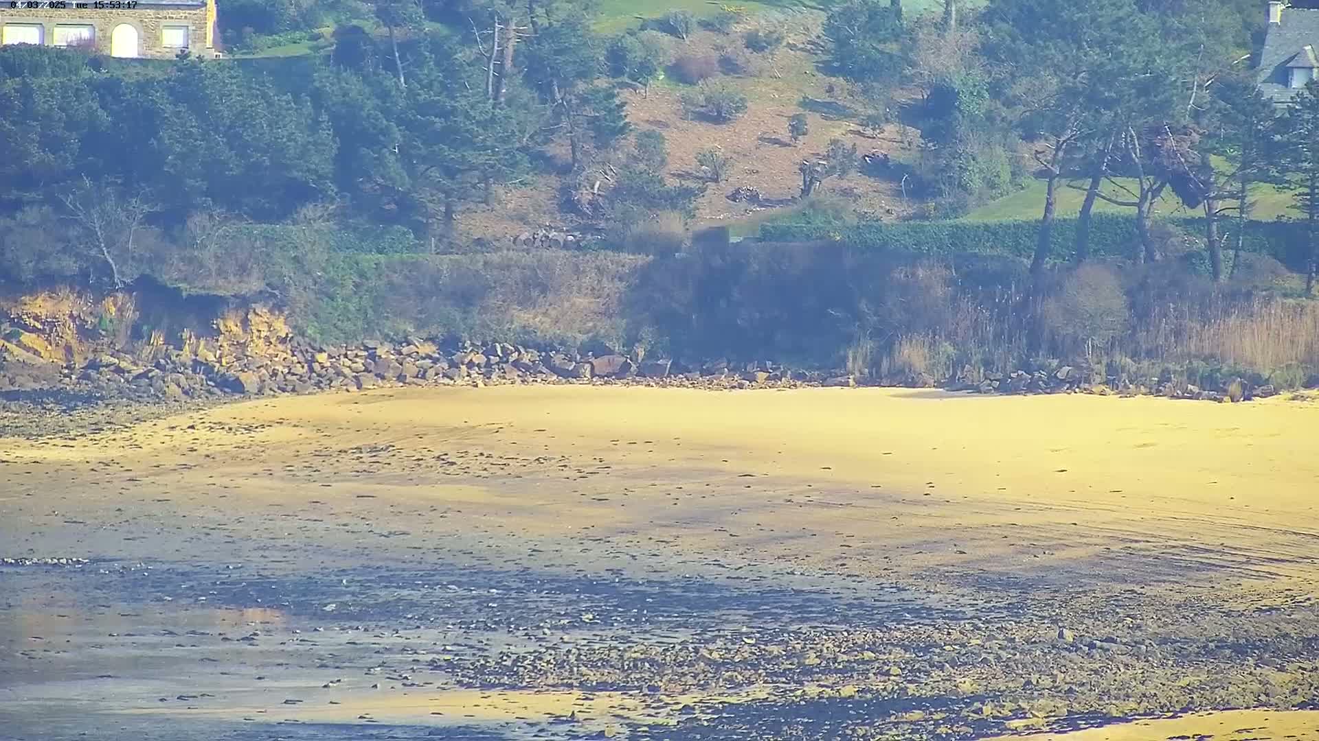 A sandy beach stretches out to a rocky shoreline, backed by a treeline on a hillside overlooking the water under a bright, sunny sky.