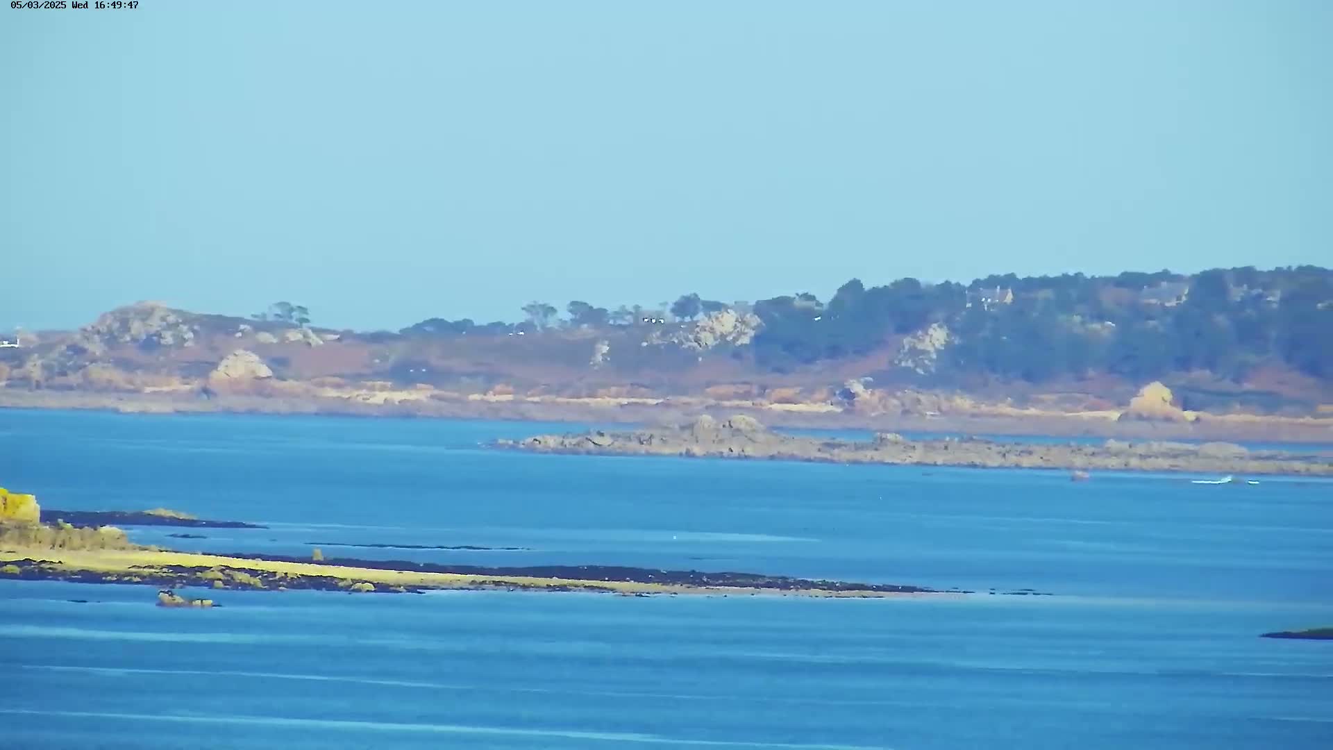 A tranquil coastal scene shows a calm blue sea with dark rocky shorelines and a distant tree-lined landmass under a clear sky.