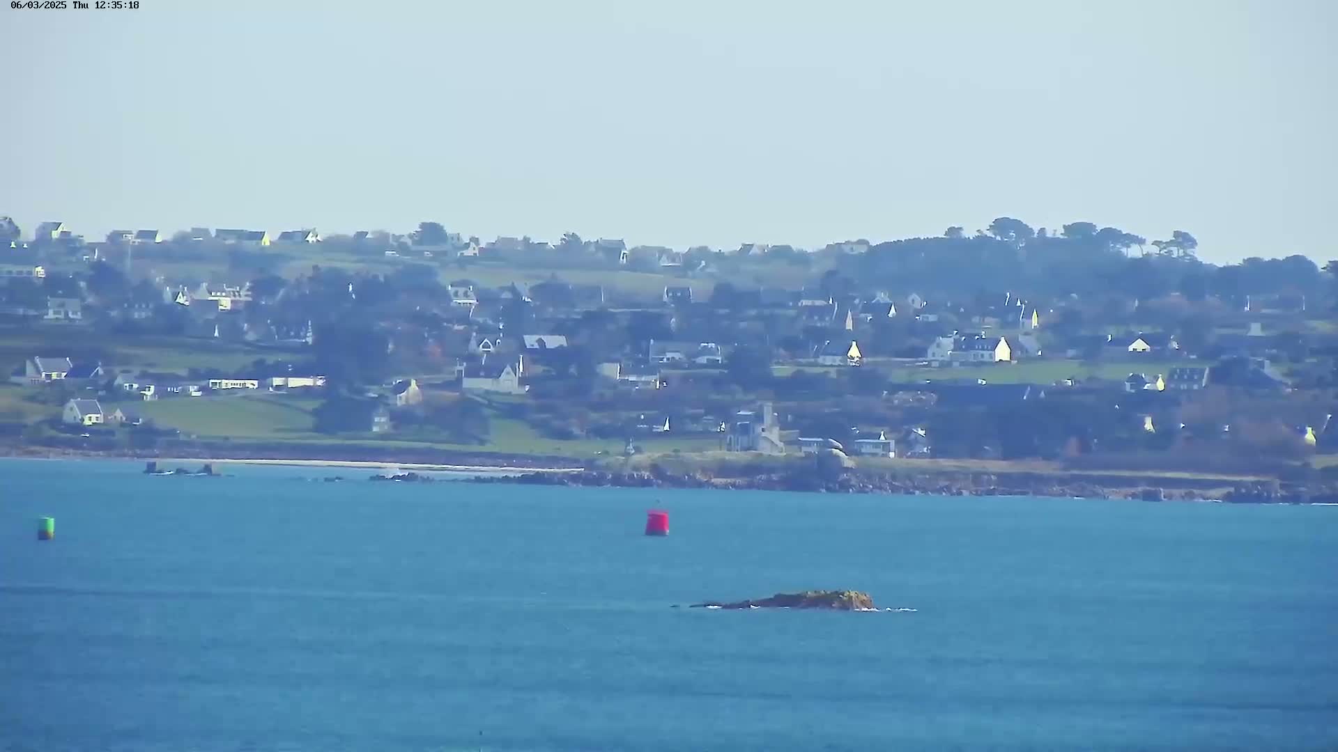 A coastal town is visible across a calm, blue body of water, with a few small islands and buoys in the foreground.