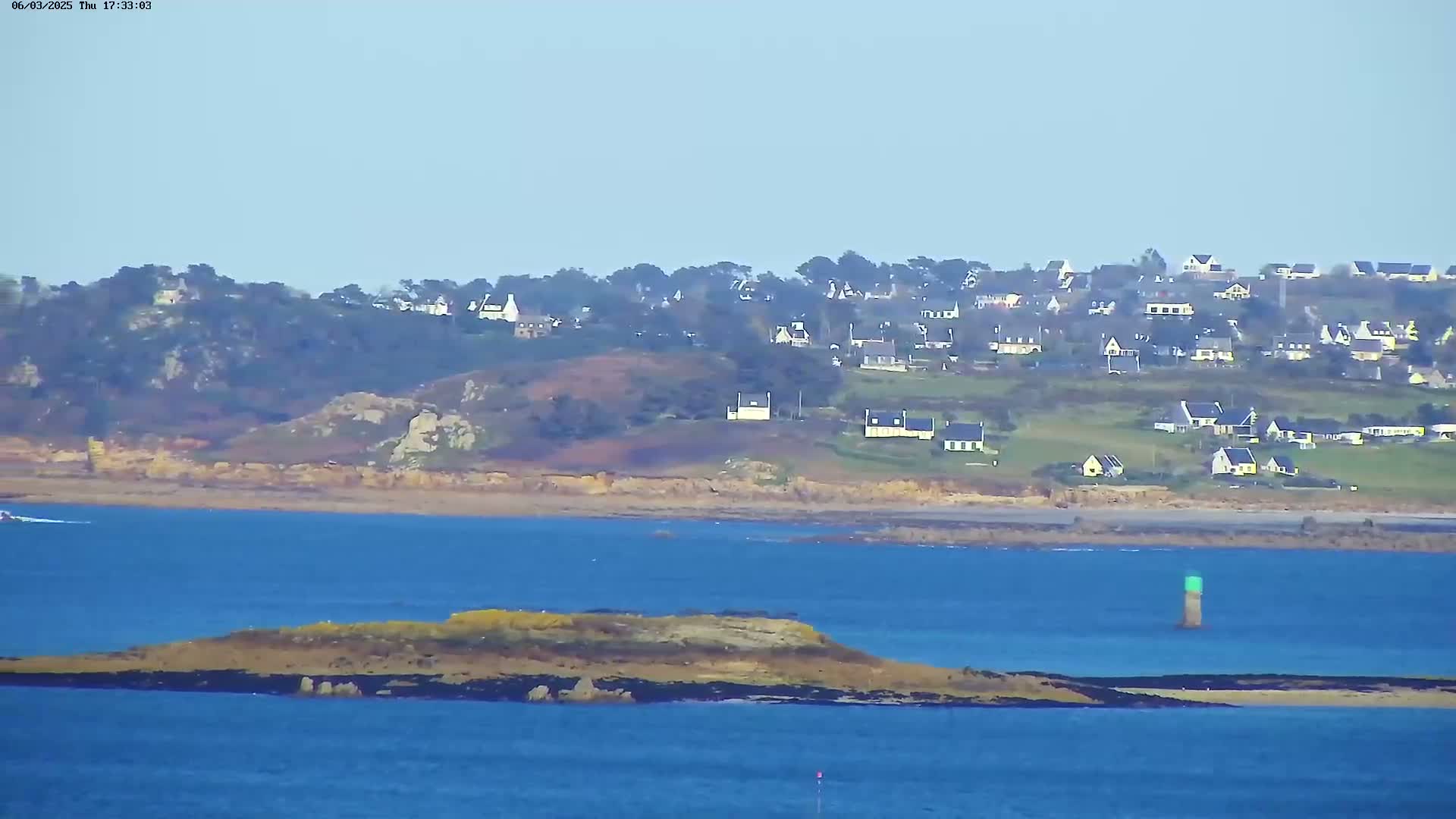A coastal scene on a clear day shows a village on a hillside overlooking a calm body of water with a small island in the foreground.