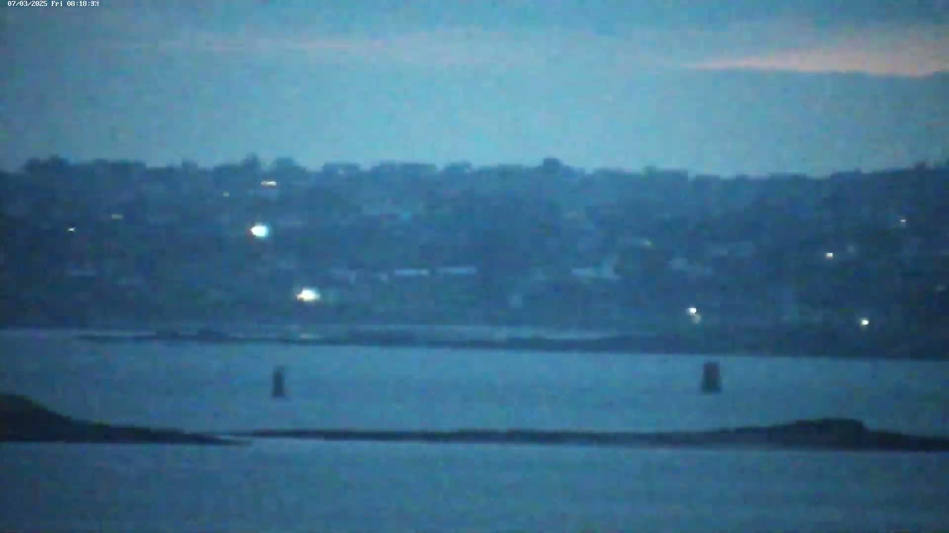 A dark, calm body of water stretches out in front of a distant, dimly lit town under a dusky sky.