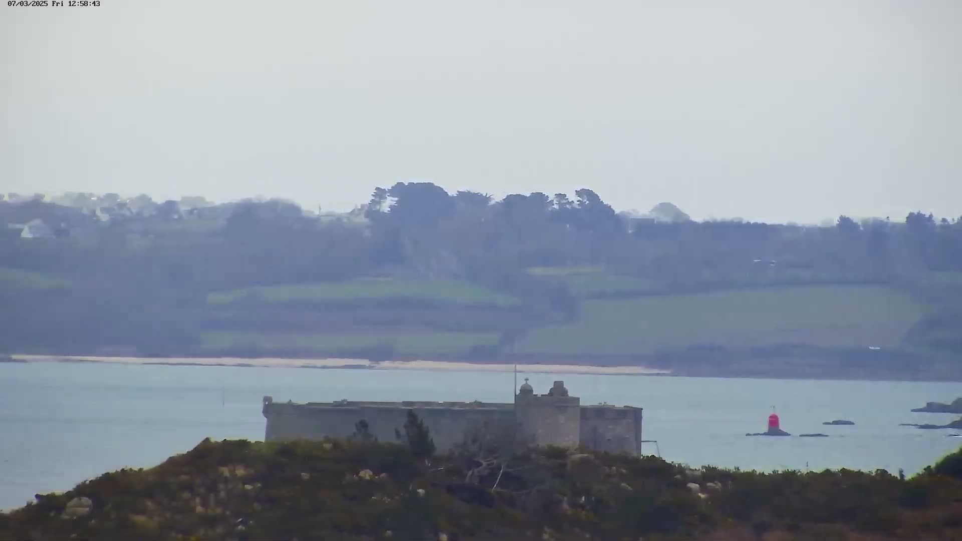 A stone fortress sits on a small, low-lying island in a calm body of water, with hazy hills and greenery in the background under an overcast sky.