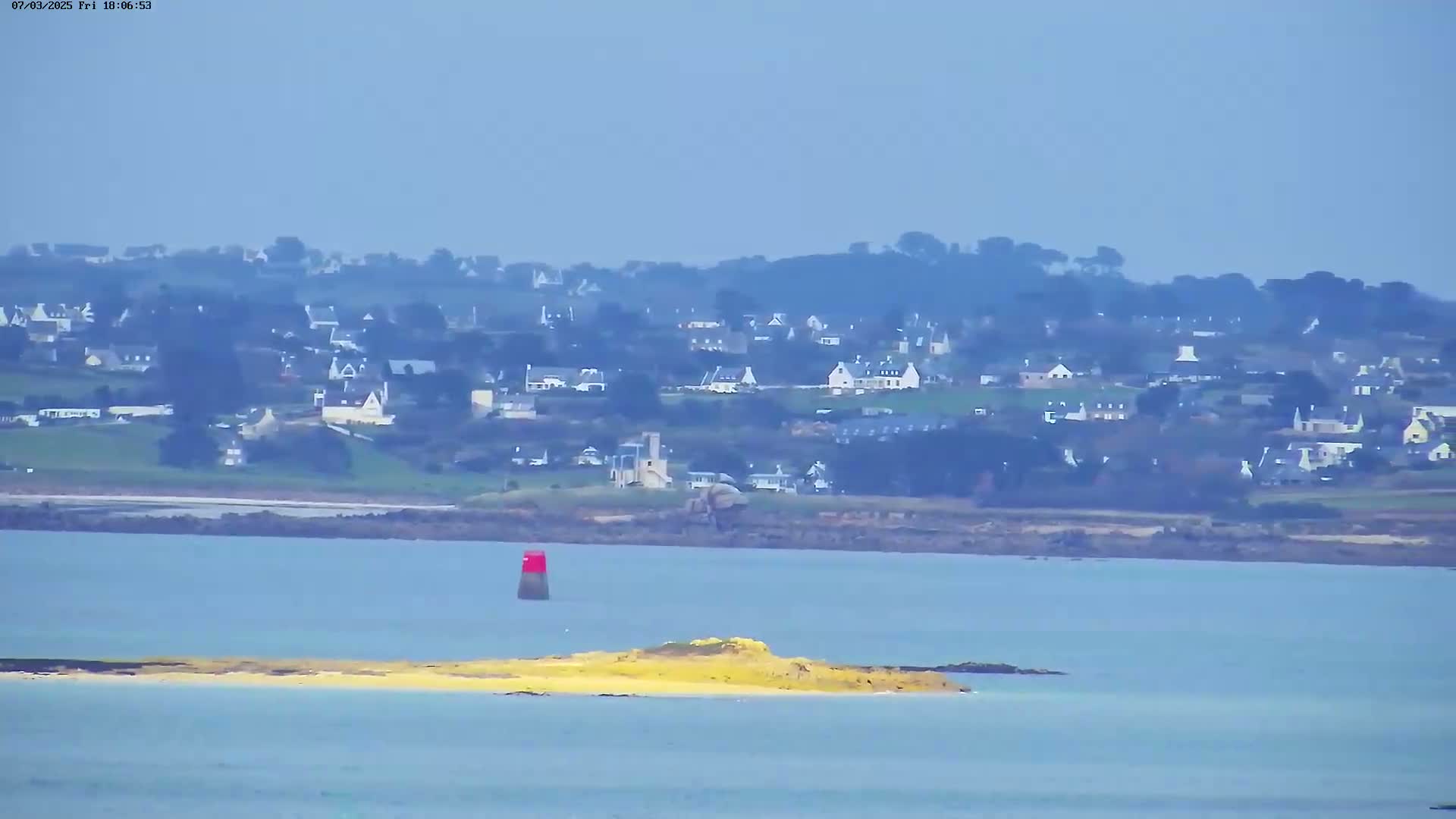 A hazy view shows a coastal town across a body of water, with a small island and a red navigational marker in the foreground.