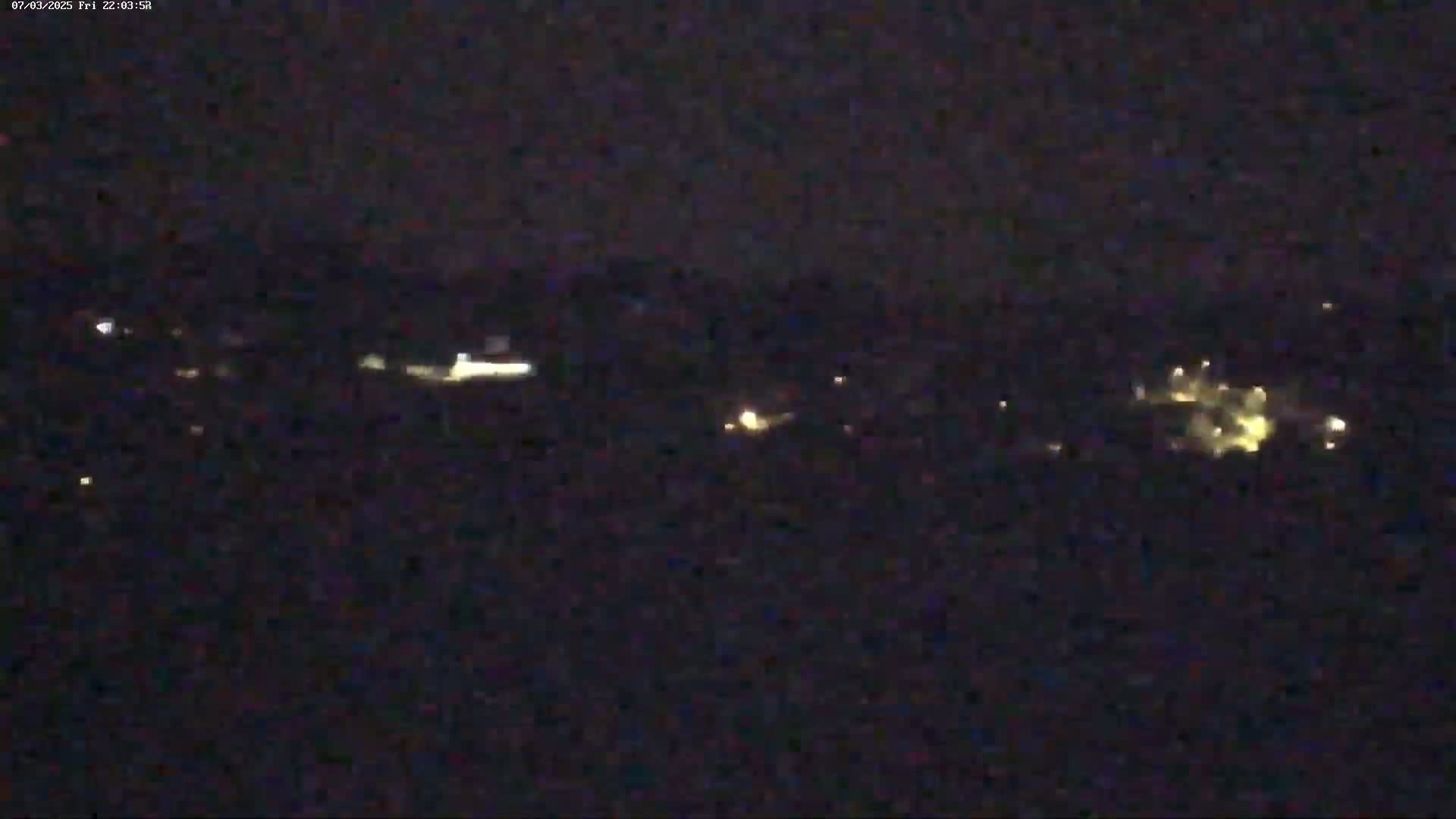 A nighttime long shot shows a dark landscape with scattered small, faint lights in the distance under a clear, dark sky.