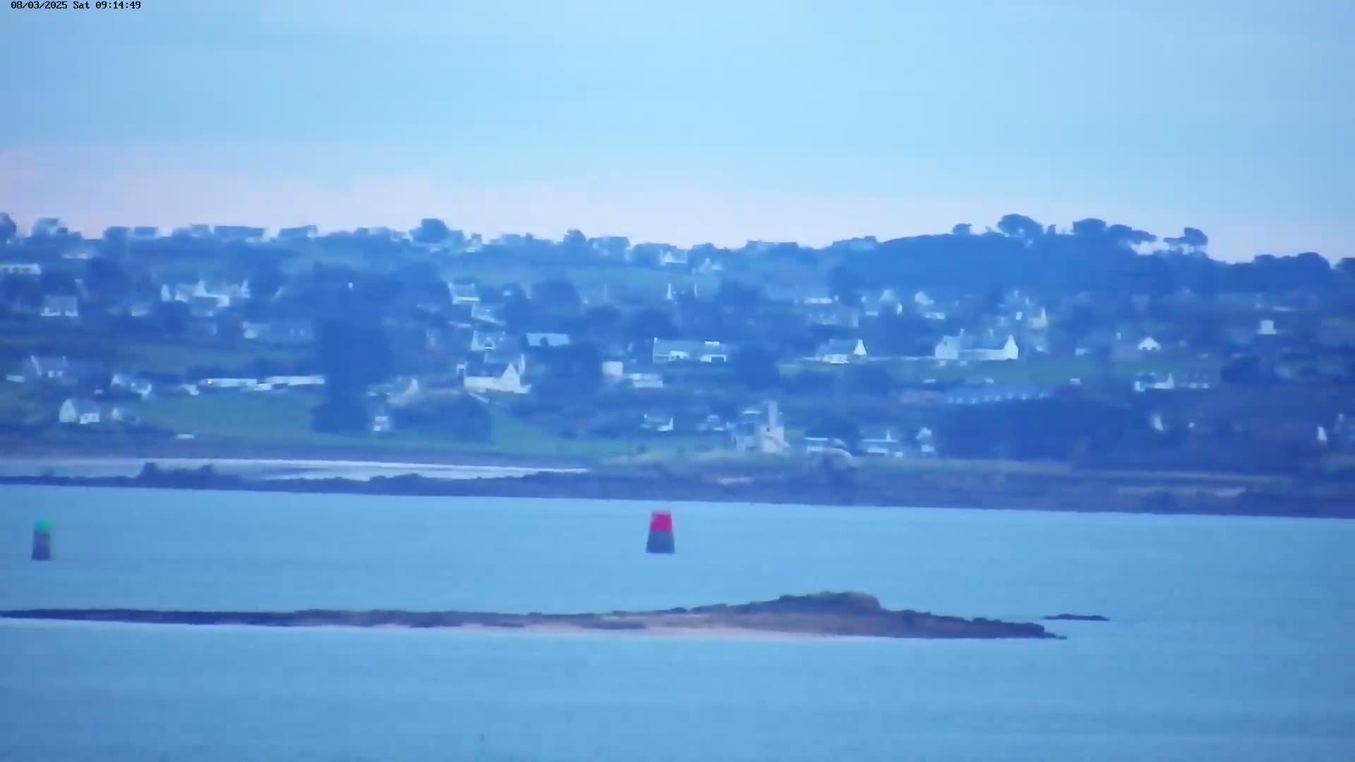 A hazy coastal scene shows a village nestled on a hillside overlooking calm blue water, marked by buoys, and a small island.