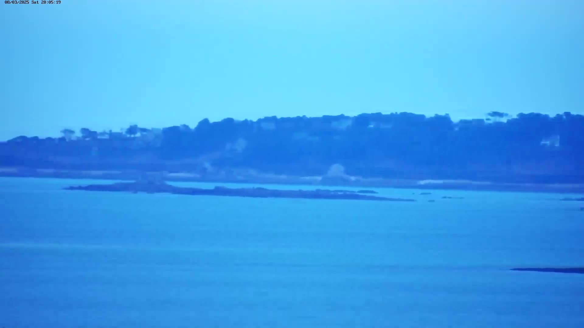 A tranquil body of water stretches to a distant shoreline of dark-green trees under a clear, light-blue sky.
