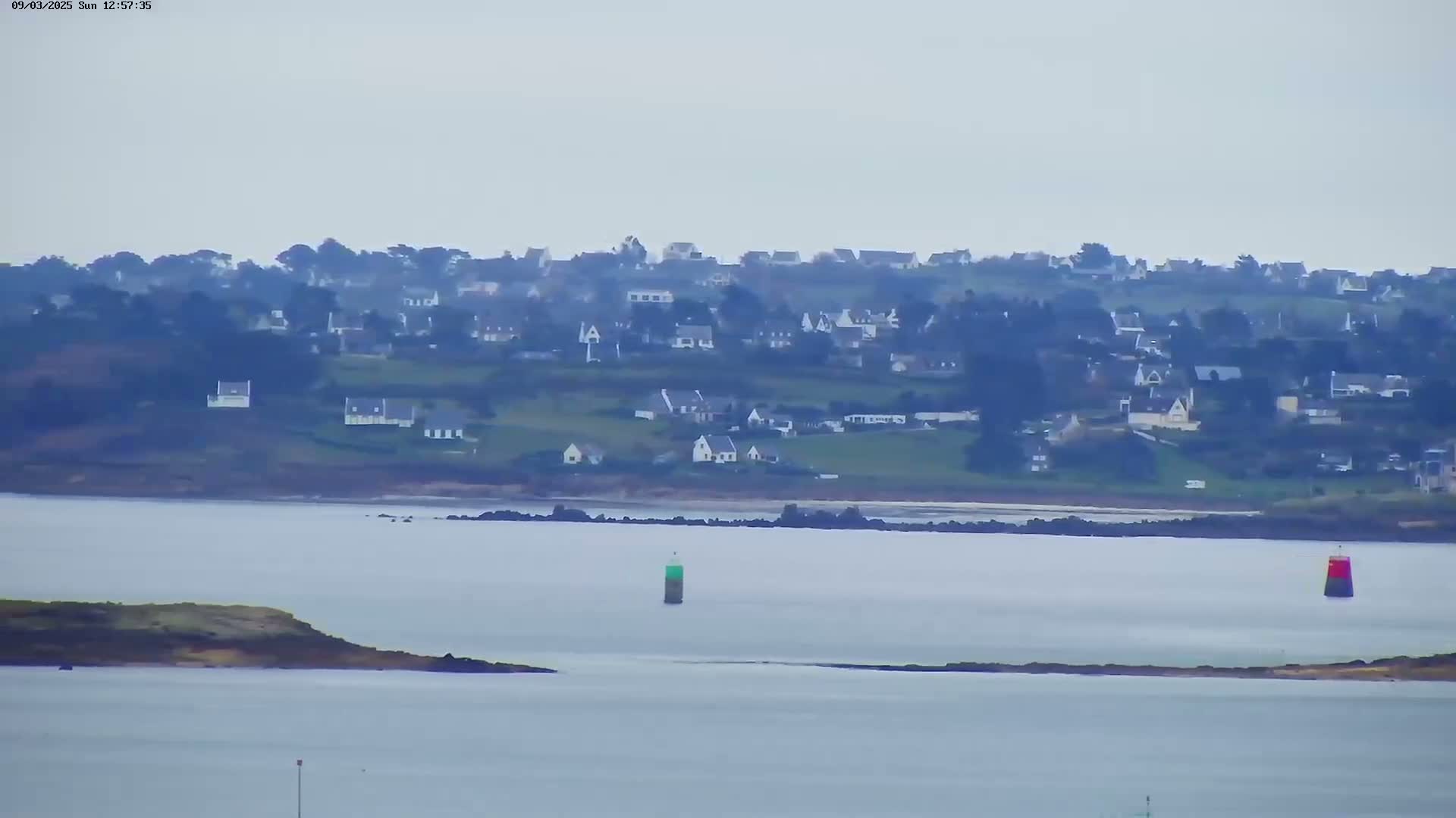 A hazy view shows a coastal village nestled on a hillside overlooking calm water with two buoys visible in the bay.