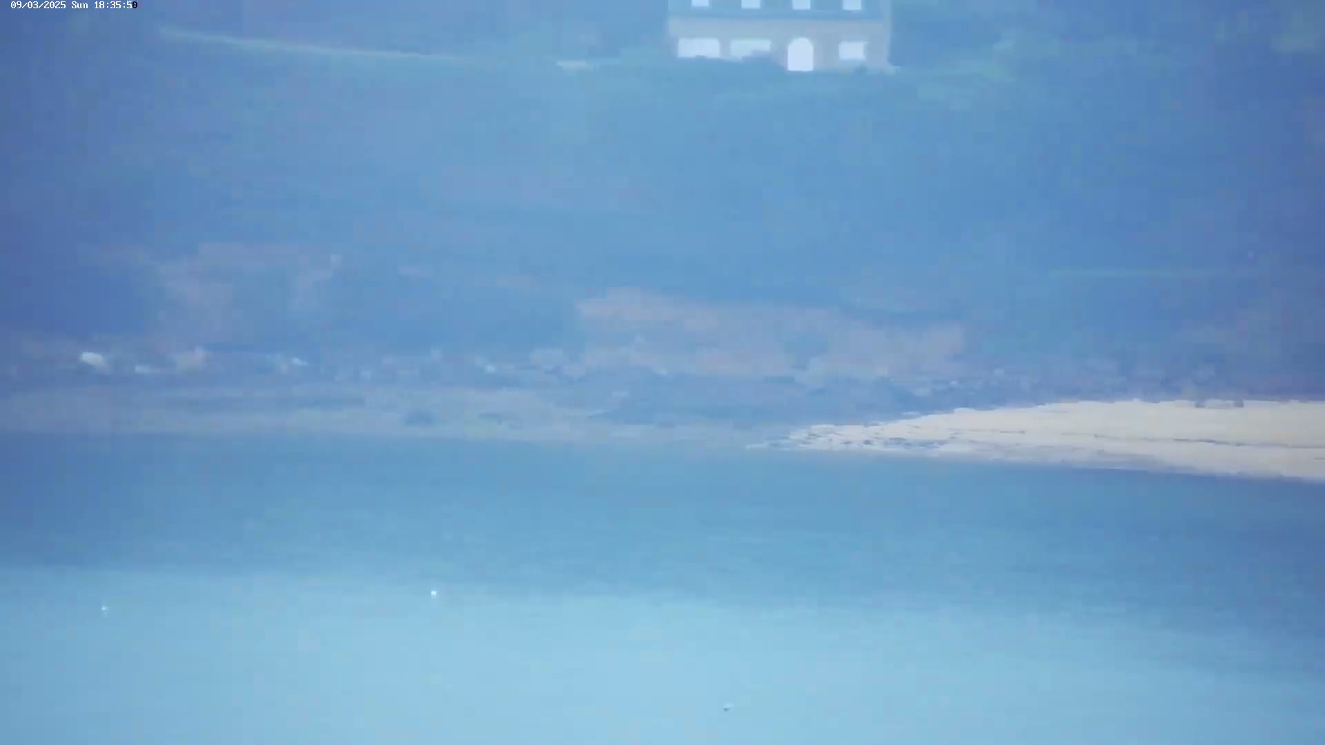 A hazy, distant view shows a light-colored beach and calm water, with a house and greenery visible in the background.