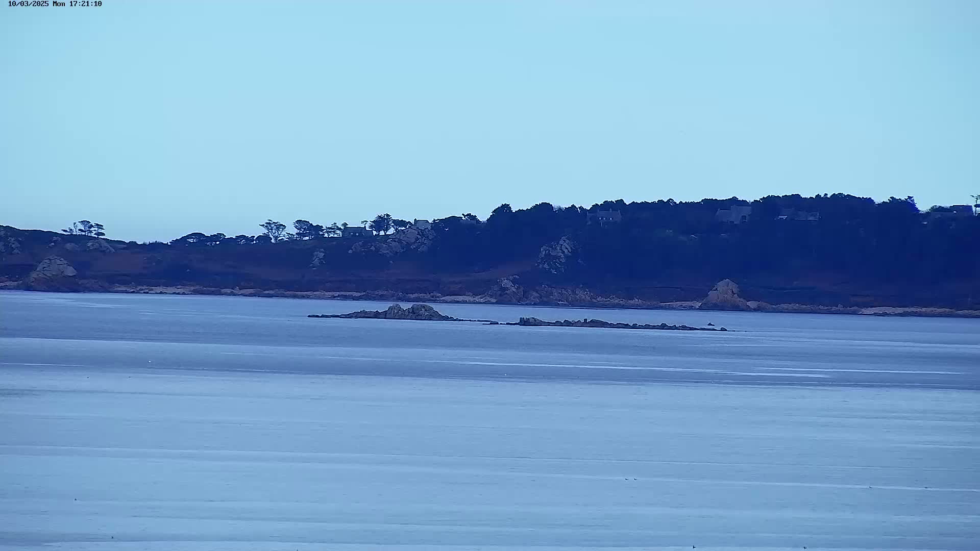 A calm, light-blue body of water stretches out before a dark, tree-covered shoreline with several rocky outcrops in the foreground.