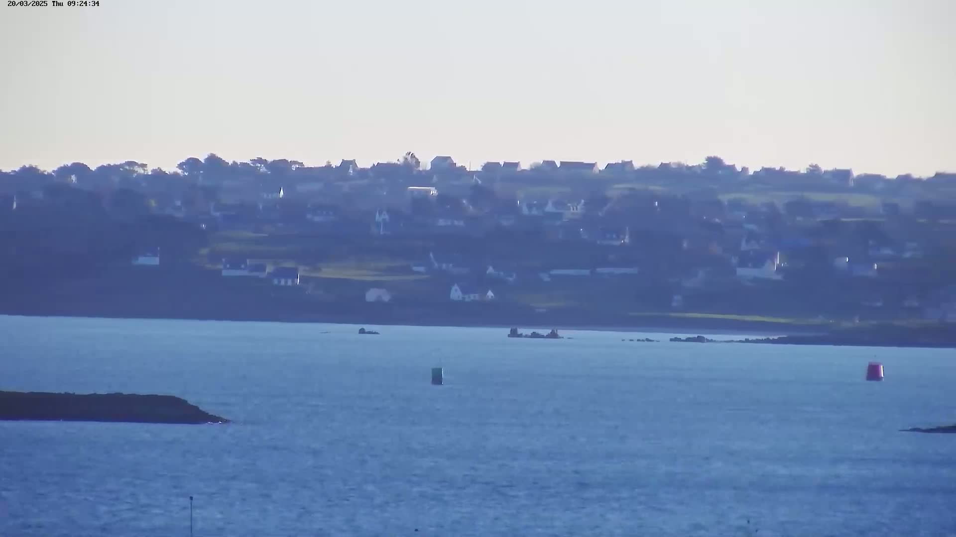 A distant coastal town is visible across a calm body of water under a hazy sky.