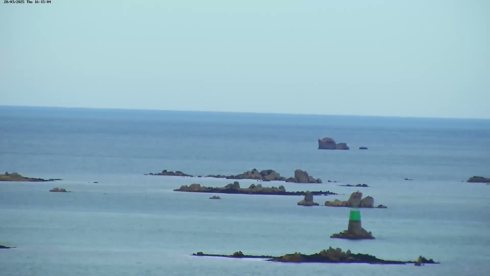 A calm, light-blue ocean stretches to the horizon, dotted with numerous dark rocky outcrops and a single green navigational buoy.