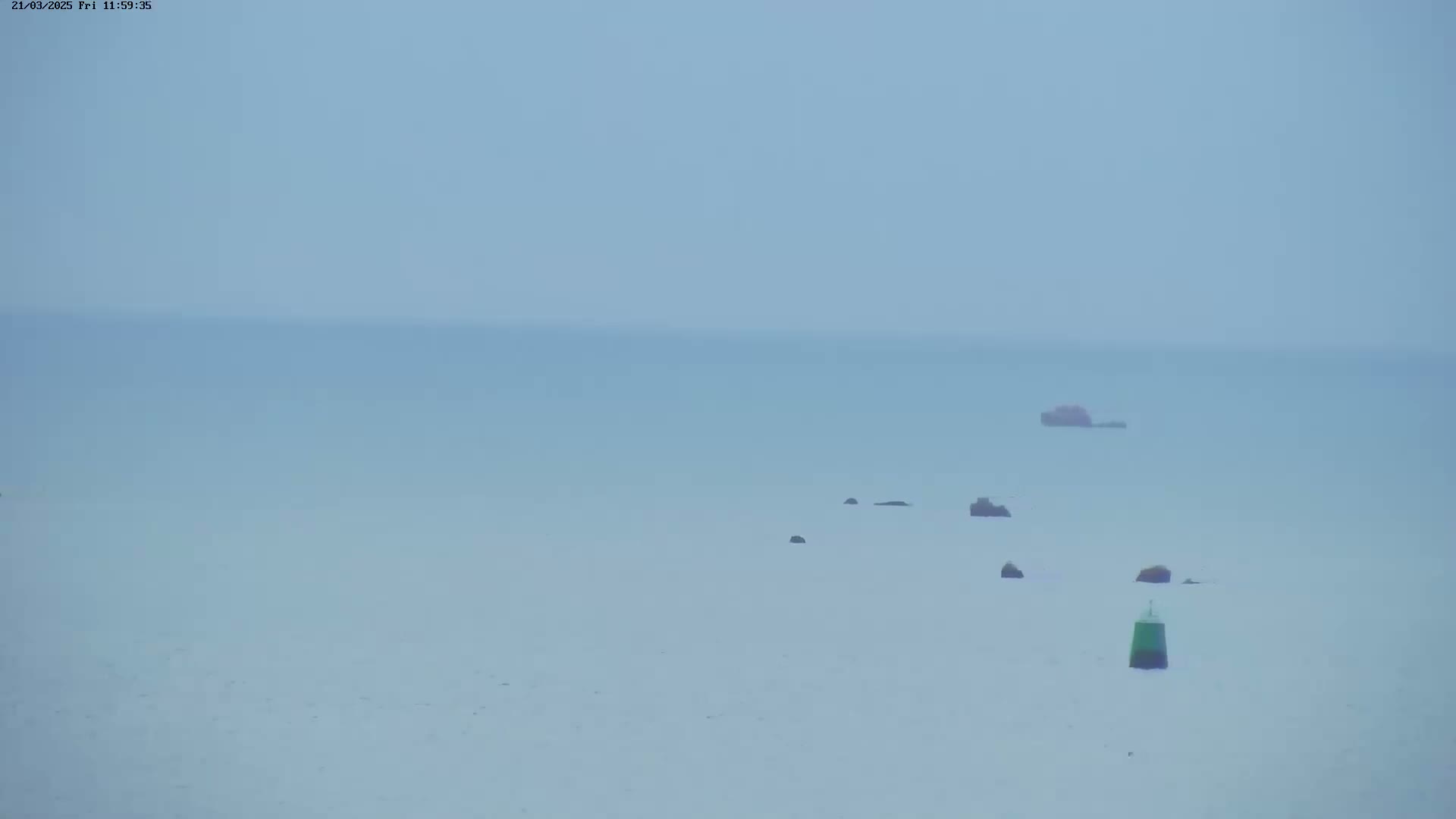A hazy, light-blue sea stretches to the horizon, featuring several dark rocks and a single green buoy.