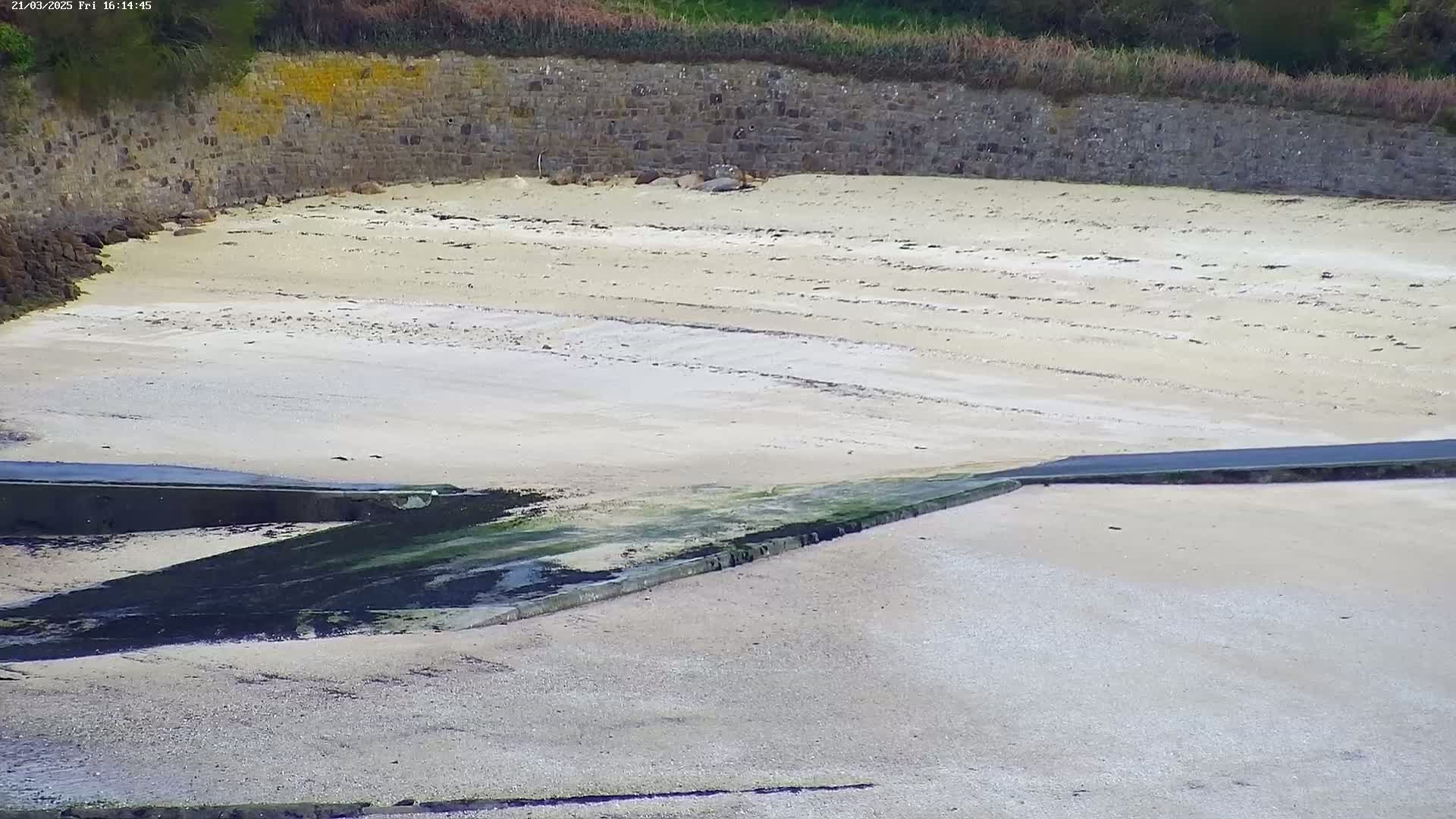 A sandy beach with a stone wall in the background and a dark-colored ramp leading into the water is shown.
