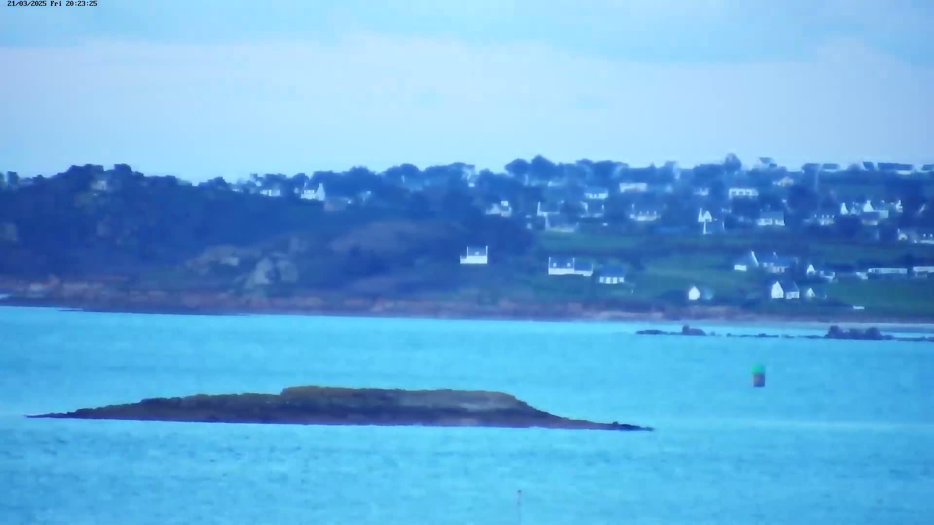 A small, dark island sits in calm, blue water in front of a distant hillside town under an overcast sky.