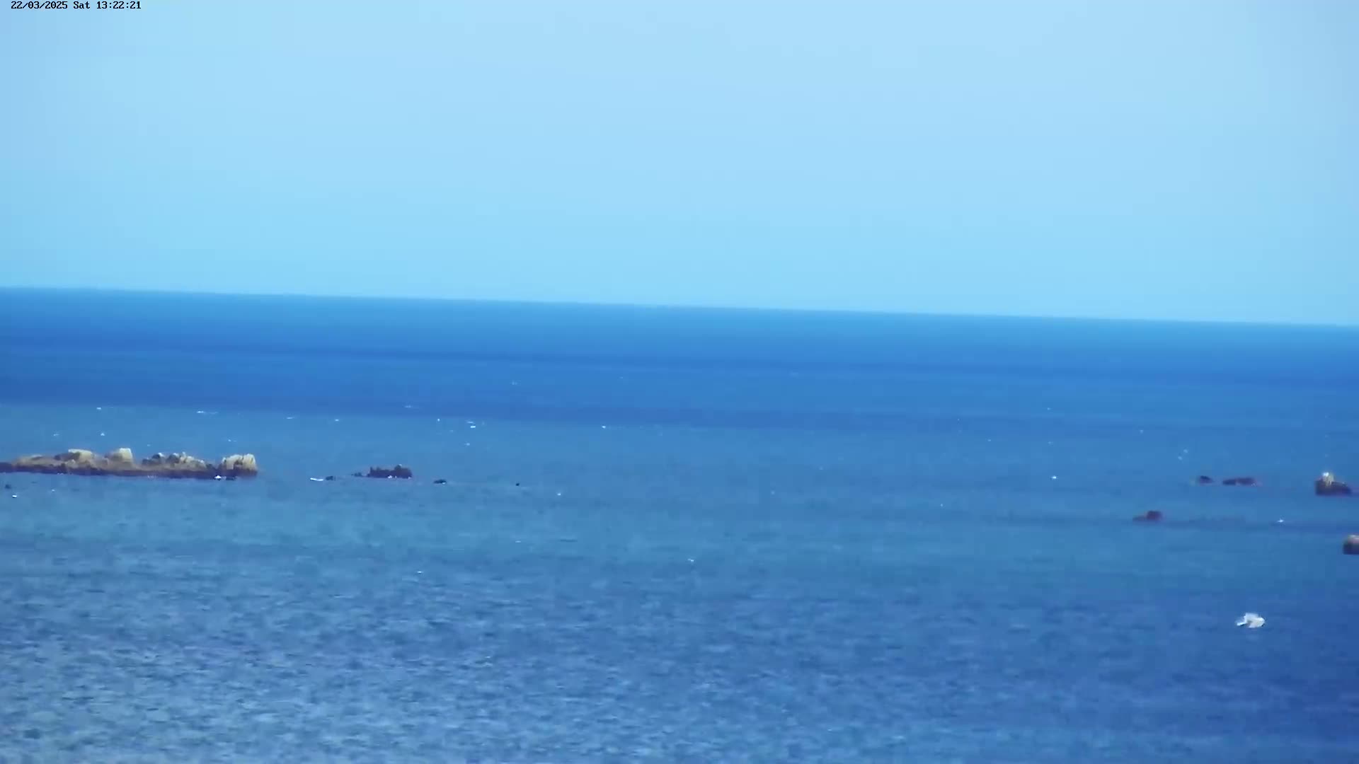 A calm, blue ocean with several small rocky islands or outcroppings visible on the horizon under a clear sky.