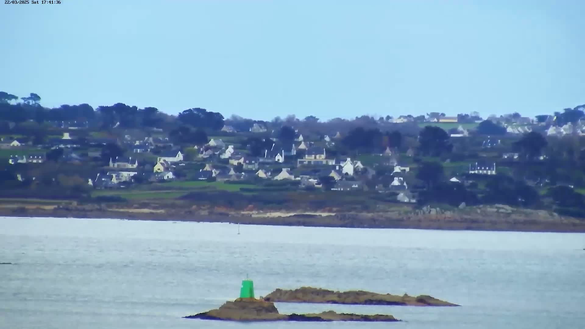A small coastal village sits on a hillside overlooking a calm body of water with a few small rocky islands and a green buoy visible in the foreground under a clear blue sky.