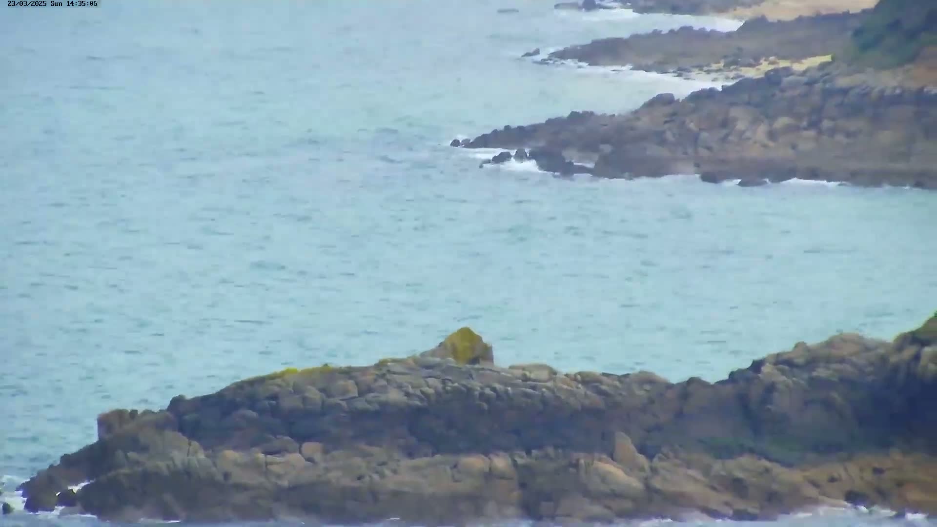 Dark, rocky coastline meets a light blue, somewhat choppy sea under a cloudy sky.