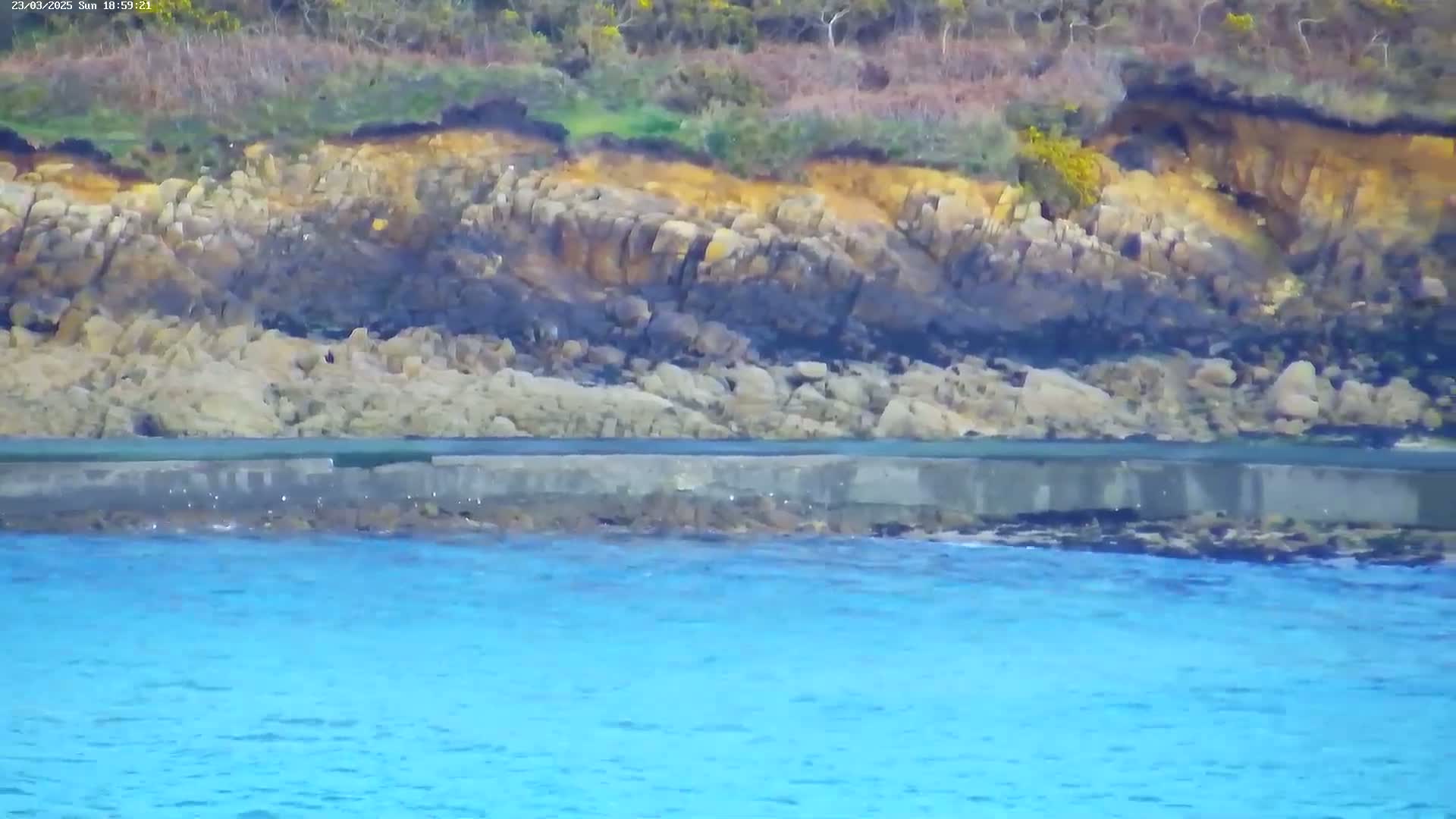 A rocky shoreline with varying shades of yellow, brown, and black rocks meets a calm, bright blue body of water.