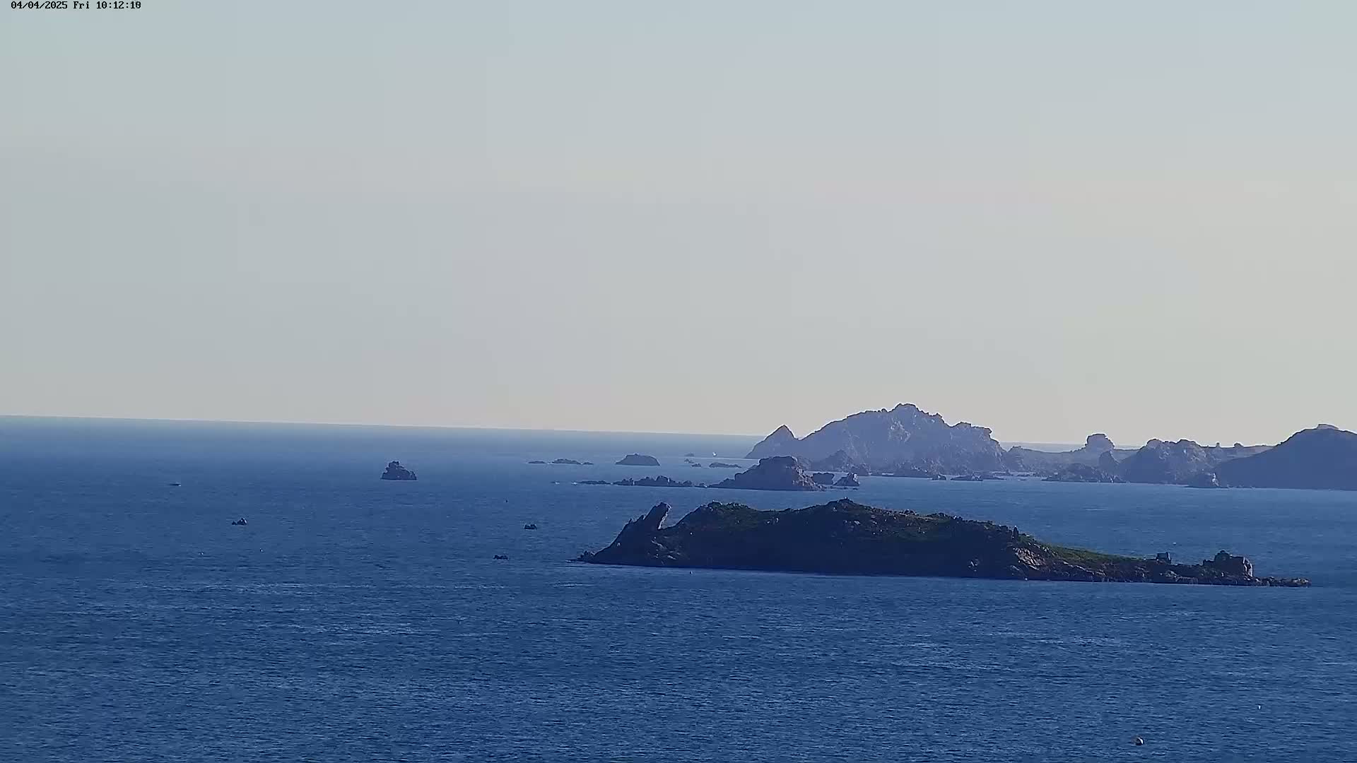 A calm, blue ocean stretches to a hazy horizon, dotted with small islands and rocky outcrops under a clear sky.