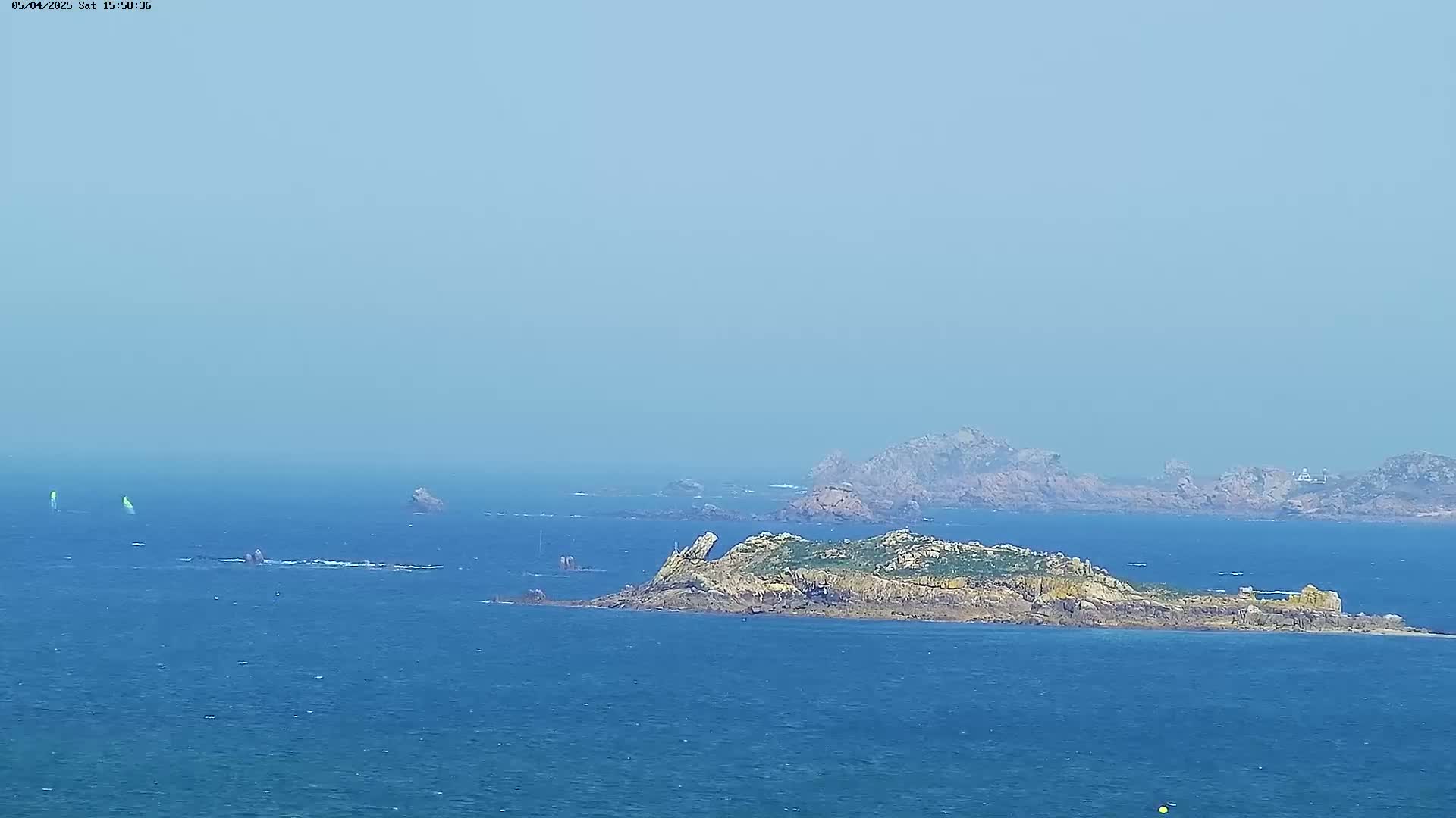 A hazy, blue day shows a rocky island in a calm, blue sea with a few sailboats in the distance.