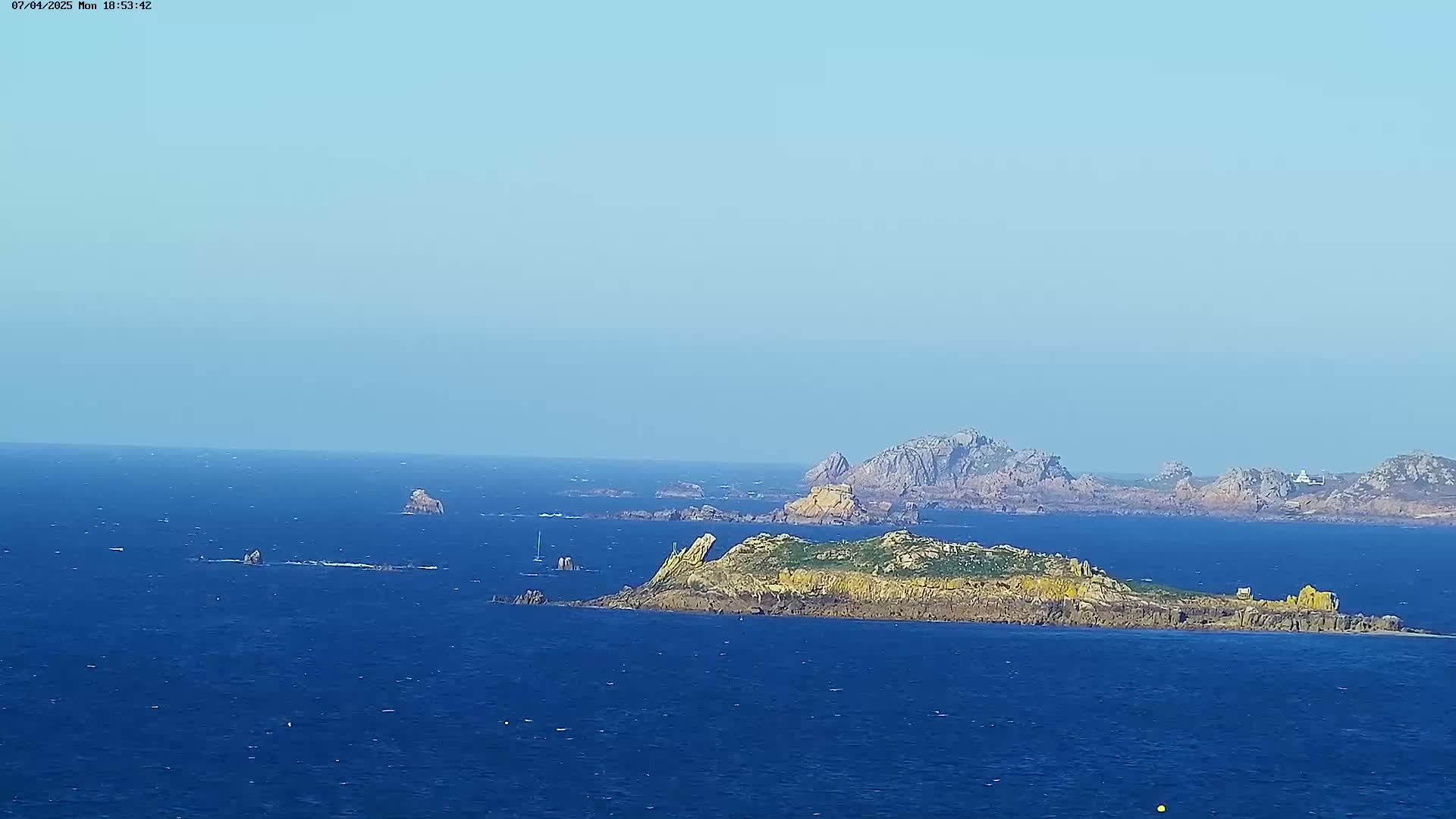 A picturesque coastal scene on a clear day shows a rocky island in the foreground, with other islands and landmasses visible in the distance across a calm, blue sea.