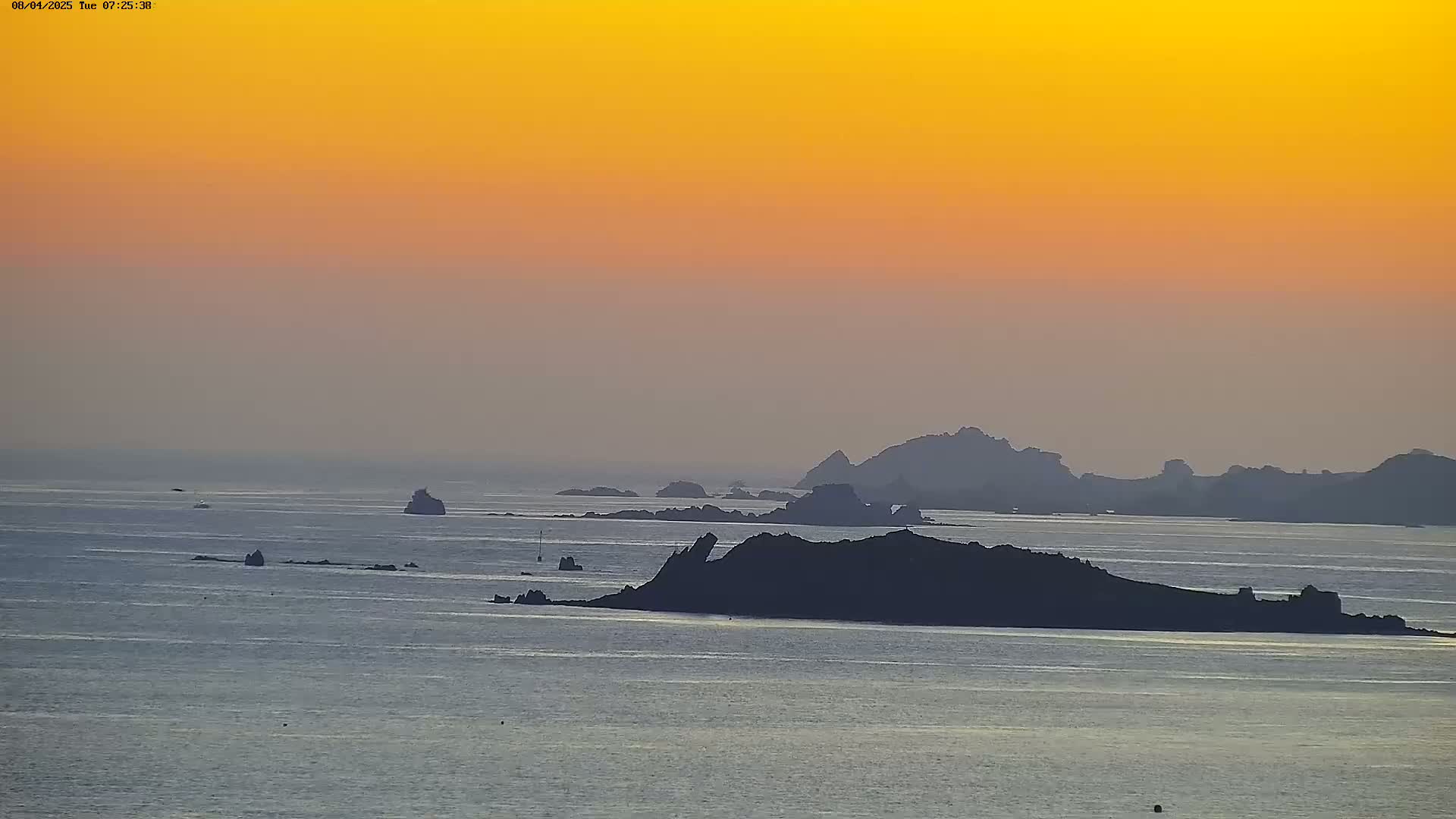 A calm ocean with several dark, rocky islands silhouetted against a vibrant orange sunset sky.