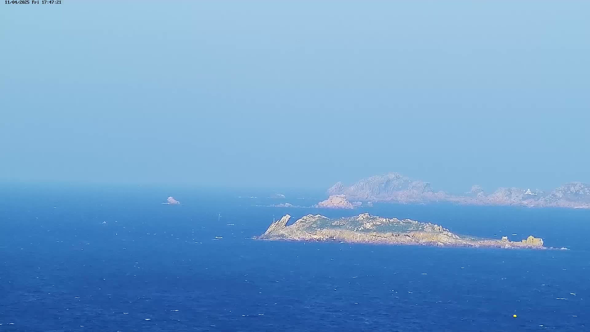 A rocky island with low-lying vegetation sits in a calm, blue ocean under a hazy sky.