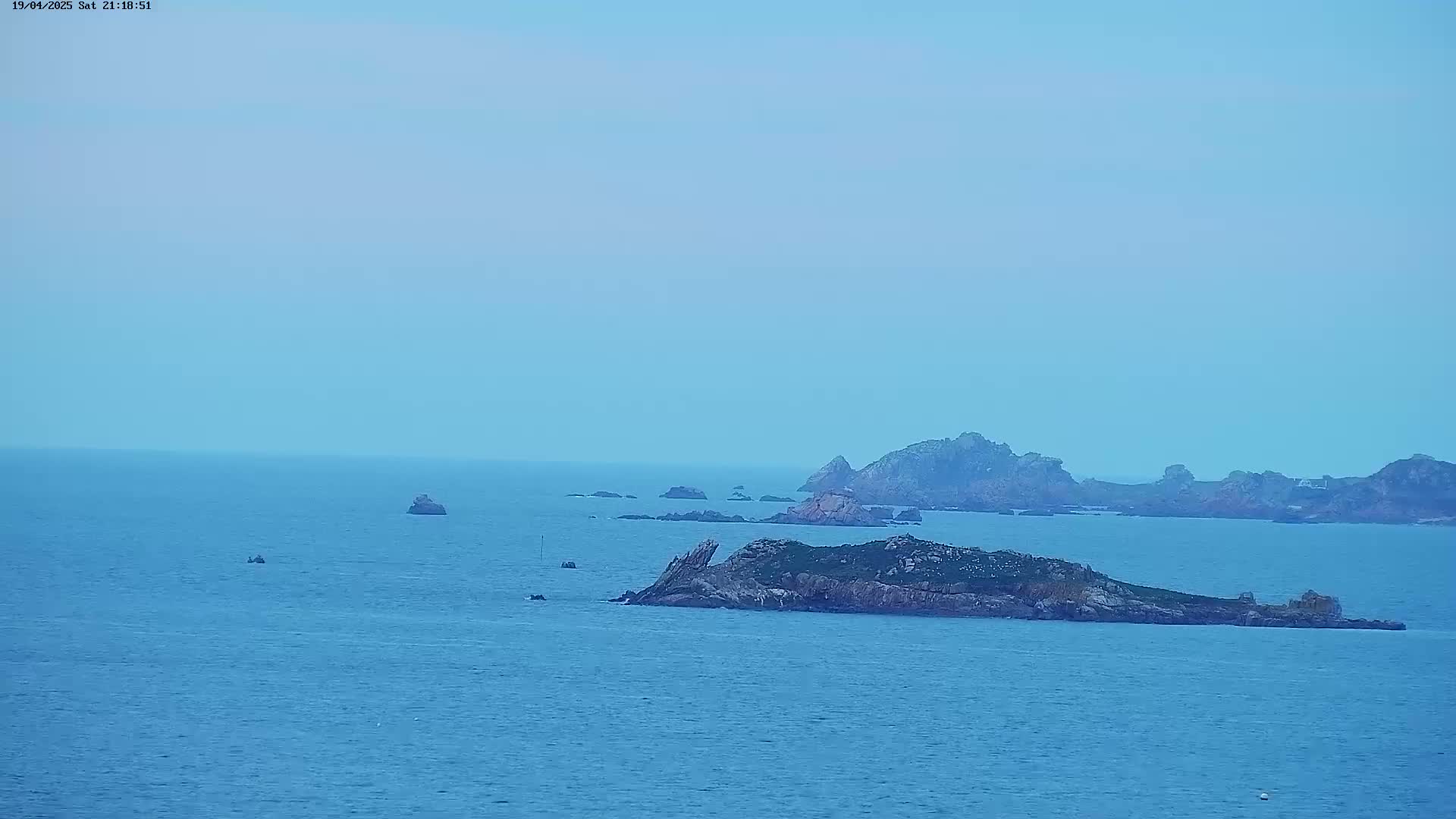 A calm, blue ocean stretches to the horizon, dotted with several small, rocky islands under a pale blue sky.