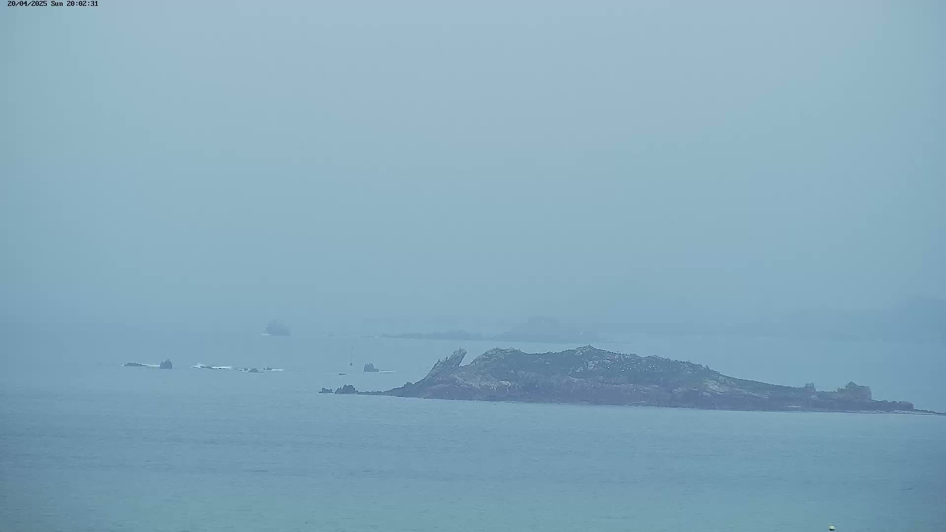 A hazy, distant view of a rocky island and smaller outcroppings in a calm sea.