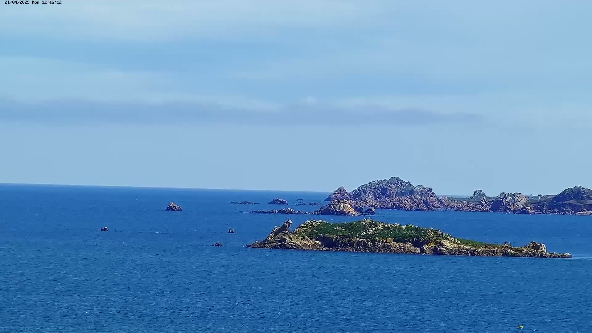 A small, vegetated island and several rocky outcrops are visible in a calm, blue ocean under a clear, light-blue sky.