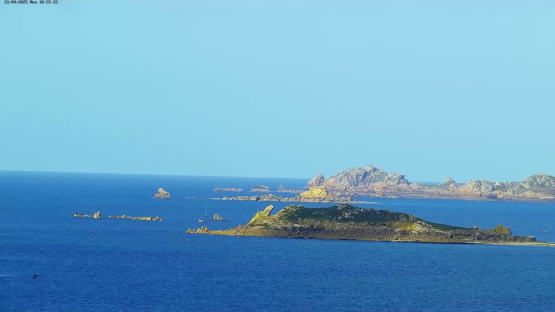 A clear, sunny day reveals a rocky coastline with numerous small islands and outcrops surrounded by a calm, blue sea.