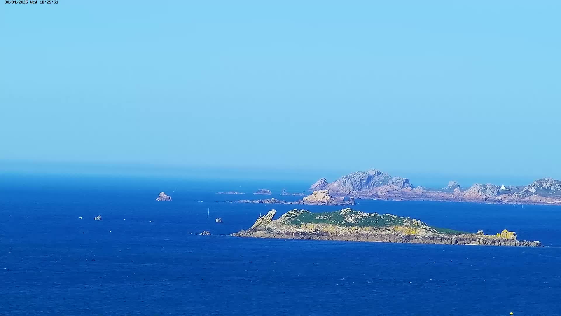 A clear, sunny day reveals a rocky island in a vast blue ocean, with other distant islands and landmasses visible on the horizon.