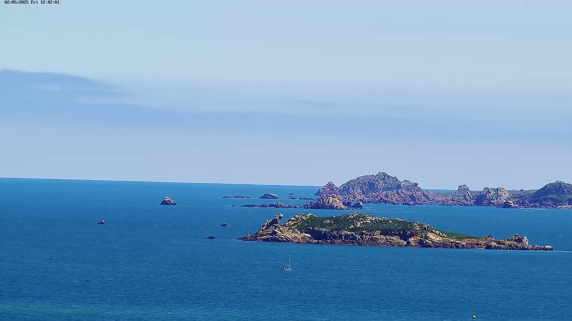 A sailboat is visible on a calm, blue ocean with several rocky islands under a mostly clear sky.