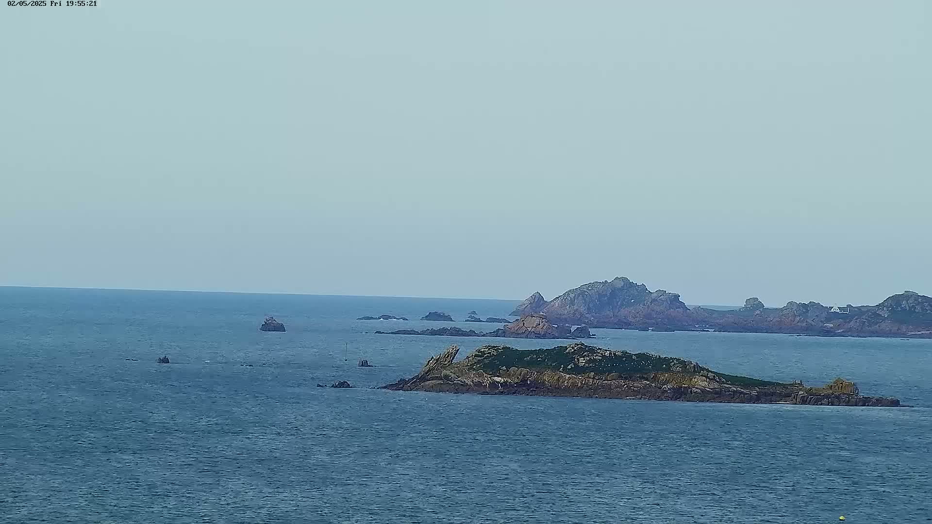 A calm, overcast day reveals a rocky island in the foreground, with other similar islands and outcrops scattered across a blue sea in the distance.